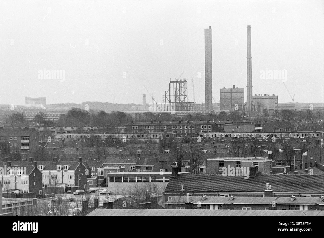 Übersicht Beverwijk Velsen Noord Overviews, Whizgle News, Dutch Desk, Niederlande, 1950 - 2000 am 18-04-1972. Das Bild enthält diese Themen. Die Szene entfaltet sich in einer Industrielandschaft, die durch eine gedämpfte Palette von Grau- und Brauntönen gekennzeichnet ist. Im Vordergrund fügt sich eine Ansammlung flacher Gebäude mit schrägen Dächern in den Blick ein, deren Strukturen eng zusammengefügt sind. Viele dieser Gebäude weisen ein verwittertes Erscheinungsbild auf, was auf eine lange Geschichte hindeutet, wobei gelegentlich Anzeichen von Verschleiß an den Fassaden zu sehen sind. Wenn sich das Auge in den Hintergrund bewegt, entsteht ein starker Kontrast mit aufragendem Rauch Stockfoto