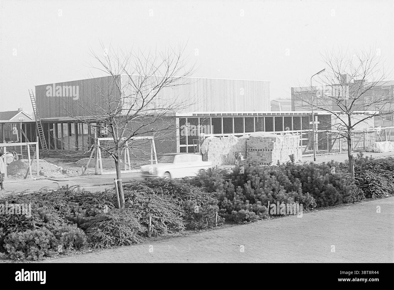 Zusätzliches Clubgebäude Rijsenhout Exterior, Whizgle News, Dutch Desk, Niederlande, 1950 - 2000 am 19.12.1968. Diese Themen sind in der Abbildung dargestellt. Die Szene zeigt eine Baustelle mit einer teilweise gebauten Struktur, die den Vordergrund dominiert. Das Gebäude hat ein modernes Design, das durch eine Kombination aus Holzlatten und großen Glaspaneelen gekennzeichnet ist, die eine luftige, zeitgenössische Ästhetik vermitteln. Es scheint eine einstöckige Struktur zu sein, und der Rahmen weist auf bedeutende Fortschritte in Bereichen hin, die noch exponiert sind. Um das Gebäude herum befinden sich mehrere Stapel von Baumaterialien, einschließlich Stockfoto