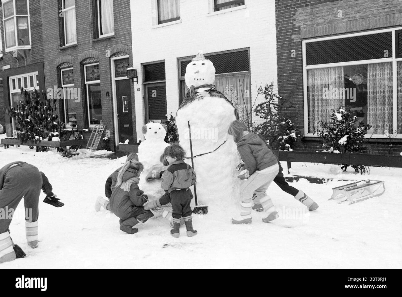 Kinder mit großem Schneemann., Whizgle News, Dutch Desk, Niederlande, 1950 - 2000. Die Abbildung zeigt diese Themen. In einer verschneiten urbanen Szene versammelt sich eine Gruppe von Kindern aufgeregt vor einer Reihe von Backsteinhäusern. Der Boden ist mit einer dicken Schneedecke bedeckt, dämpft Geräusche und schafft ein winterliches Wunderland. Ein Schneemann, größer als die Kinder, steht stolz in der Mitte. Dieser Schneemann ist mit einem provisorischen Schal und einem rudimentären Gesichtsausdruck verziert, mit dunklen Gegenständen, die Augen und einen Mund andeuten. Die Kinder, gebündelt in farbenfroher Winterkleidung, zeigen eine Reihe von lebhaften Farben – Re Stockfoto Kinder mit großem Schneemann., Whizgle News, Dutch Desk, Niederlande, 1950 - 2000. Die Abbildung zeigt diese Themen. In einer verschneiten urbanen Szene versammelt sich eine Gruppe von Kindern aufgeregt vor einer Reihe von Backsteinhäusern. Der Boden ist mit einer dicken Schneedecke bedeckt, dämpft Geräusche und schafft ein winterliches Wunderland. Ein Schneemann, größer als die Kinder, steht stolz in der Mitte. Dieser Schneemann ist mit einem provisorischen Schal und einem rudimentären Gesichtsausdruck verziert, mit dunklen Gegenständen, die Augen und einen Mund andeuten. Die Kinder, gebündelt in farbenfroher Winterkleidung, zeigen eine Reihe von lebhaften Farben – Re Stockfoto