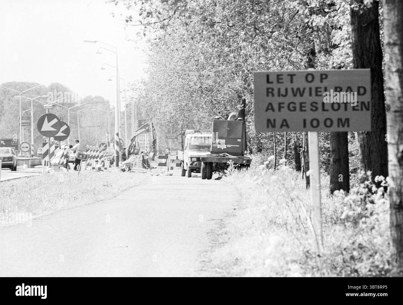 Radweg Arbeit Amsterdamseweg IJmuiden Radwege Radfahrer arbeiten IJmuiden Amsterdamseweg Niederlande, Whizgle News, Dutch Desk, Niederlande, 1950 - 2000 am 14-05-1980. Diese Themen werden in der Abbildung angezeigt. Die Szene zeigt eine Straße entlang einer belebten Baustelle. Der Asphalt wirkt verschlissen und gedämpft, was zu der Distanz führt, in der er sich windet. Auf beiden Seiten bilden Bäume mit üppig grünem Laub eine natürliche Grenze, deren Blätter von Sonnenlicht durchdrungen sind und Schatten auf die Straße werfen. Ein kreisförmiges Verkehrsschild steht gut sichtbar und führt die Fahrzeuge vorsichtig herum Stockfoto