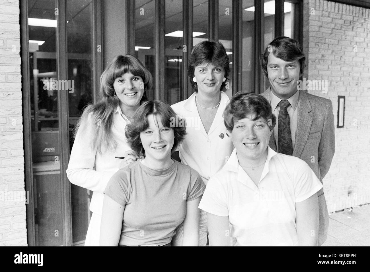Ein Team von fünf Personen bei der Arbeit, einer im Büro, einer hinter der Theke und zusammen vor dem Eingang., Whizgle News, Dutch Desk, Niederlande, 1950 - 2000 am 06-1980. Die Abbildung zeigt diese Themen. In der Szene versammelt sich eine Gruppe von fünf Personen, die ein Gefühl von Kameradschaft und Wärme ausstrahlen. Der Hintergrund ist mit einer hellen Ziegelwand versehen, die durch große Fenster verstärkt wird, die weiches, diffuses natürliches Licht eindringen lassen und so eine sanfte Atmosphäre schaffen. Die erste Reihe besteht aus drei Frauen mit jeweils unterschiedlichen Frisuren. Die linke Figur hat langes, welliges Haar. Stockfoto