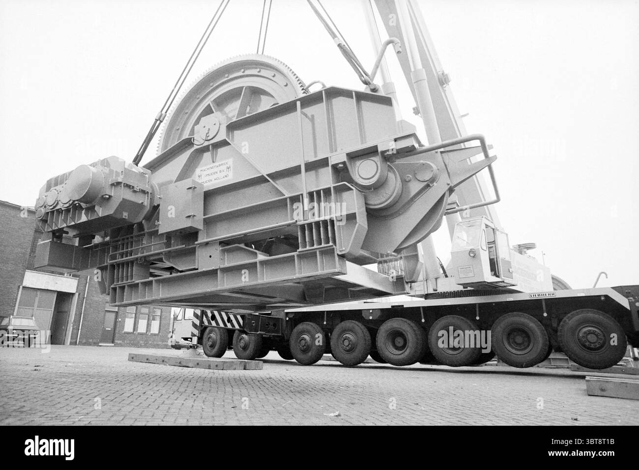 Entladung großer Windenmaschinenfabrik IJmuiden Maschinen und Fabriken IJmuiden Velsen IJmuiden Niederlande, Whizgle News, Dutch Desk, Niederlande, 1950 - 2000 am 06-10-1981. Dies sind die Themen im Bild. In einer geschäftigen industriellen Umgebung steht ein großes, schweres Maschinenteil im Mittelpunkt, das an einem Kran in der Luft hängt. Die Maschine verfügt über einen robusten, rechteckigen Körper mit verschiedenen mechanischen Komponenten, einschließlich Zahnrädern und einem markanten kreisförmigen Rad auf einer Seite. Die Oberfläche ist in einem gedämpften Grauton lackiert, was ein Gefühl von Festigkeit und Haltbarkeit vermittelt. Um diesen Ingenieur herum Stockfoto