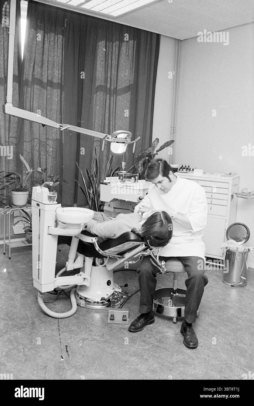 Zahnzentrum Innenzahnärzte, Whizgle News, Dutch Desk, Niederlande, 1950 - 2000 am 14-03-1968. Dies sind die Elemente im Bild. Die Szene fängt eine Zahnarztpraxis mit einer klinischen und dennoch ruhigen Atmosphäre ein. Die Farbpalette besteht aus gedämpften Tönen, überwiegend sanften weiß- und Grautönen, im Kontrast zu den dunkleren Tönen der Möbel und dem tiefen Grün der großen Pflanzen im Raum. In der Mitte lehnt sich ein Zahnarztstuhl mit einem Patienten zurück. Der Patient, eine Person mit mittellangen Haaren, wirkt entspannt und liegt bequem, wenn er nach oben zeigt Stockfoto