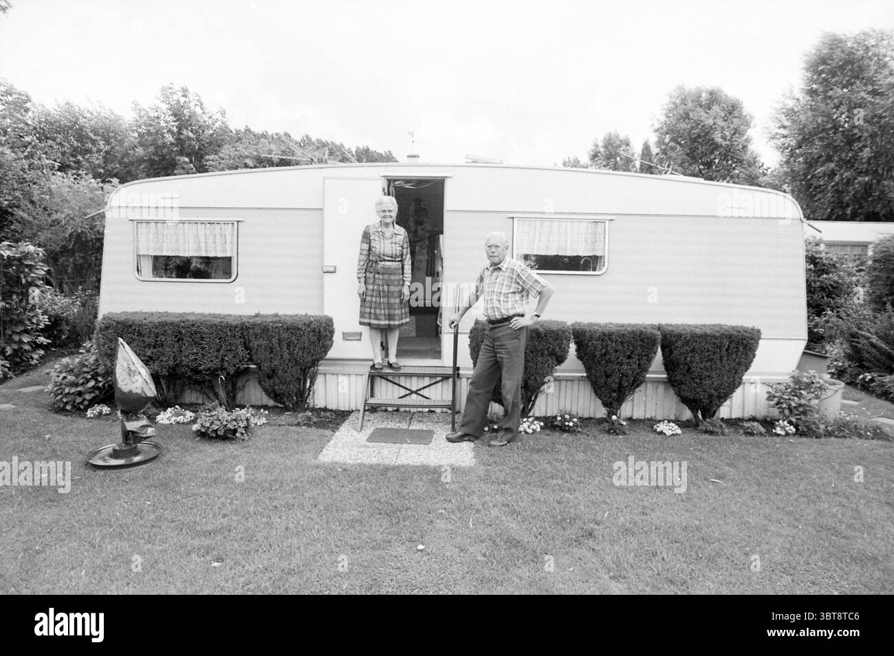 Paar vWijl Rijsenhout Rijsenhout, Whizgle News, Dutch Desk, Niederlande, 1950 - 2000 am 10-07-1990. Das Bild enthält diese Themen. Ein Paar steht vor einem bescheidenen Wohnwagen, gekennzeichnet durch seine klaren Linien und sein schlichtes Design. Der Anhänger ist in erster Linie in einer sanften, hellen Farbe gehalten und ergänzt das Grün, das ihn umgibt. Der Eingang verfügt über eine kleine Reihe von Stufen, die zu einer Tür führen, flankiert von zwei Fenstern, die mit transparenten weißen Vorhängen verziert sind, die das Sonnenlicht diffusen. Das Paar trägt zwanglose Kleidung, die für warmes Wetter typisch ist. Die Frau mit kurzem Haar trägt ein kariertes Kleid und Stockfoto