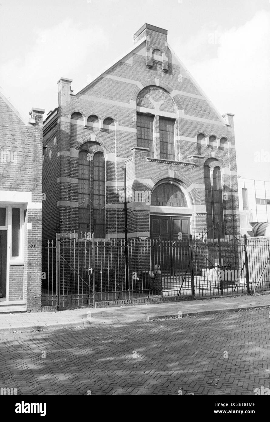 Kirche Meerplein Beverwijk Kirchen Beverwijk Meerplein Niederlande, Whizgle News, Dutch Desk, Niederlande, 1950 - 2000 am 14-10-1987. Das Bild enthält diese Themen. Die Szene zeigt ein markantes Backsteingebäude, das sich durch seinen unverwechselbaren architektonischen Stil auszeichnet. Die Struktur hat eine ausgeprägte Fassade mit komplizierten Mustern, die durch alternierende helle und dunkle Ziegelsteine geschaffen werden. Große, teilweise schattierte bogenförmige Fenster dominieren die Front und tragen zum großen Erscheinungsbild des Gebäudes bei. Der Eingang ist von einem symmetrischen Bogengang eingerahmt, der Stabilität und Stärke betont. Um das Buildin herum Stockfoto