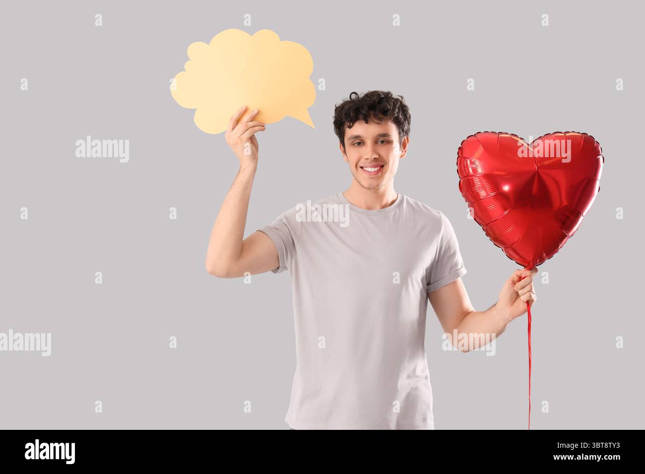 Hübscher junger glücklicher Mann mit Herzballon und Sprechblase auf grauem Hintergrund Stockfoto