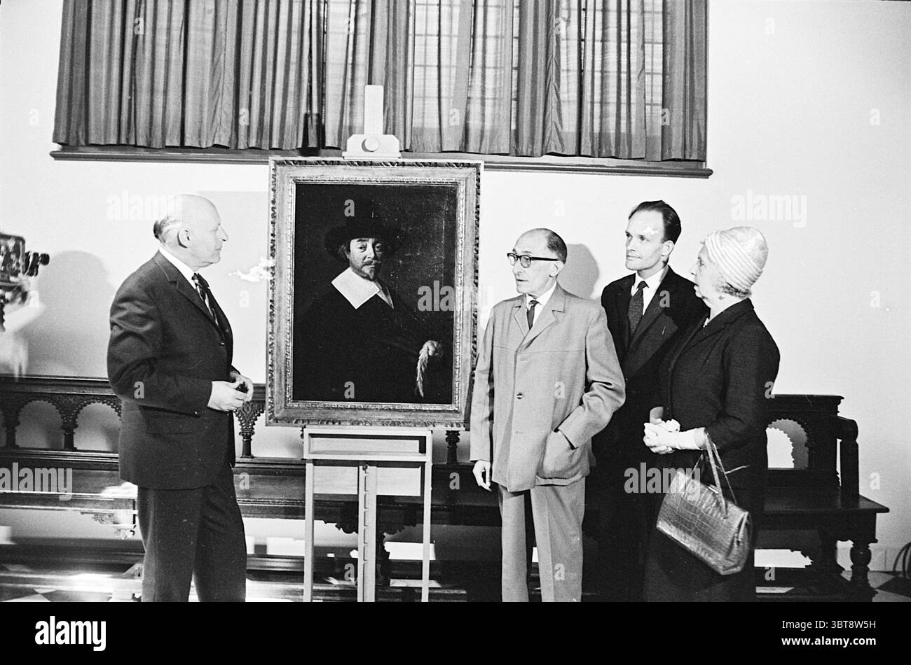 Hatinov Paar mit neu entdeckten Frans Hals Frans Hals Museum Personen, Whizgle News, Dutch Desk, Niederlande, 1950 - 2000 am 20-05-1964. Das Bild enthält diese Themen. In einem gut beleuchteten Innenbereich stehen vier Personen vor einem großen, dunkel gerahmten Gemälde, das einen Mann in Kleidung aus dem 17. Jahrhundert darstellt, komplett mit breitkrempigem Hut und Rüschen. Der Hintergrund des Gemäldes ist dunkler als die vor ihm liegenden Motive, was den ernsten Ausdruck der Figur unterstreicht. Die erste Person links, ein älterer Mann, zeigt sich auf das Gemälde und schlägt eine Diskussion ab vor Stockfoto