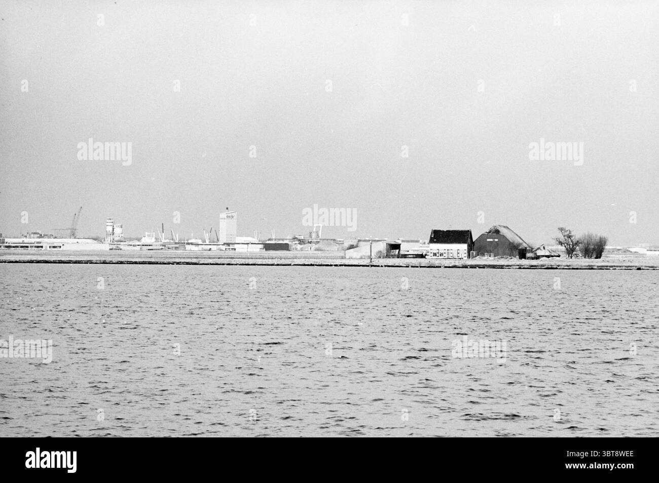 Übersicht Noordwijkermeerpolder Polder, Whizgle News, Dutch Desk, Niederlande, 1950 - 2000 am 06-01-1982. Dies sind die Elemente im Bild. Die Szene zeigt eine ruhige Uferpromenade, wo das Wasser ruhig und reflektierend ist und die subtilen Farben eines gedämpften Himmels widerspiegelt. Am Ufer kann man eine Mischung aus Gebäuden sehen – einige sind modern, mit klaren Linien und minimalistischem Stil, während andere traditionell mit schrägen Dächern sind. Der Kontrast zwischen den architektonischen Stilen sorgt für ein visuelles Interesse in der gesamten Landschaft. Die Farbpalette ist vorwiegend monochromatisch, mit Grautönen und weichen Weißtönen, W Stockfoto