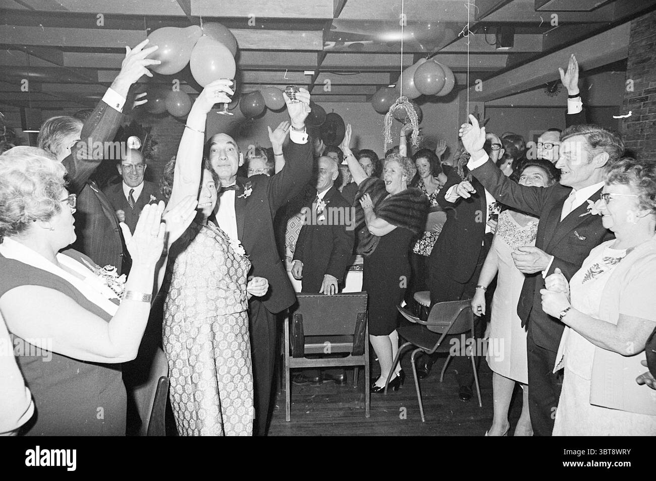 Jubilee Horsebill Jubilee Jubilee Jubiversaries, Whizgle News, Dutch Desk, Niederlande, 1950 - 2000 am 16-01-1971. Das Bild enthält diese Themen. Die Szene entfaltet sich in einer lebendigen Innenumgebung, die mit einem Gefühl von Feiern und Festlichkeiten erfüllt ist. Es sind zahlreiche Menschen versammelt, von denen jede Freude und Aufregung ausstrahlt. Viele Teilnehmer tragen formelle Kleidung, Männer tragen Anzüge und Krawatten, während Frauen elegante Kleider mit Verzierungen tragen. Farbenfrohe Ballons und Dekorationen hängen von der Decke und verleihen der Atmosphäre einen lebendigen Touch. Die Gesamtpalette verfügt über tiefe Farbtöne, die durch den br Stockfoto