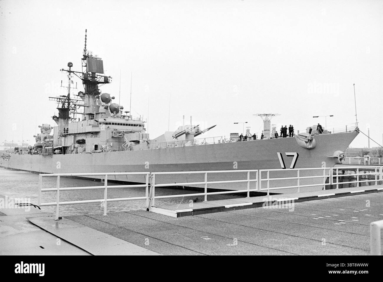 Amerikanisches Kriegsschiff in IJm. Ships, Whizgle News, Dutch Desk, Niederlande, 1950 - 2000 am 11-11-1977. Dies sind die Themen im Bild. Die Szene zeigt ein großes Marineschiff, das an einem Hafen angedockt ist. Das Schiff zeichnet sich durch seine schlanke und beeindruckende Struktur aus und ist überwiegend grau und verkörpert eine utilitaristische militärische Ästhetik. Auf dem Schiff sind verschiedene Antennen und Radargeräte zu sehen, was auf fortschrittliche technologische Fähigkeiten hindeutet. Das Schiff ist mit zahlreichen Rettungsbooten geschmückt, und mehrere Besatzungsmitglieder sind um das Deck herum positioniert, einige beobachten die Umgebung, während andere mitmachen Stockfoto