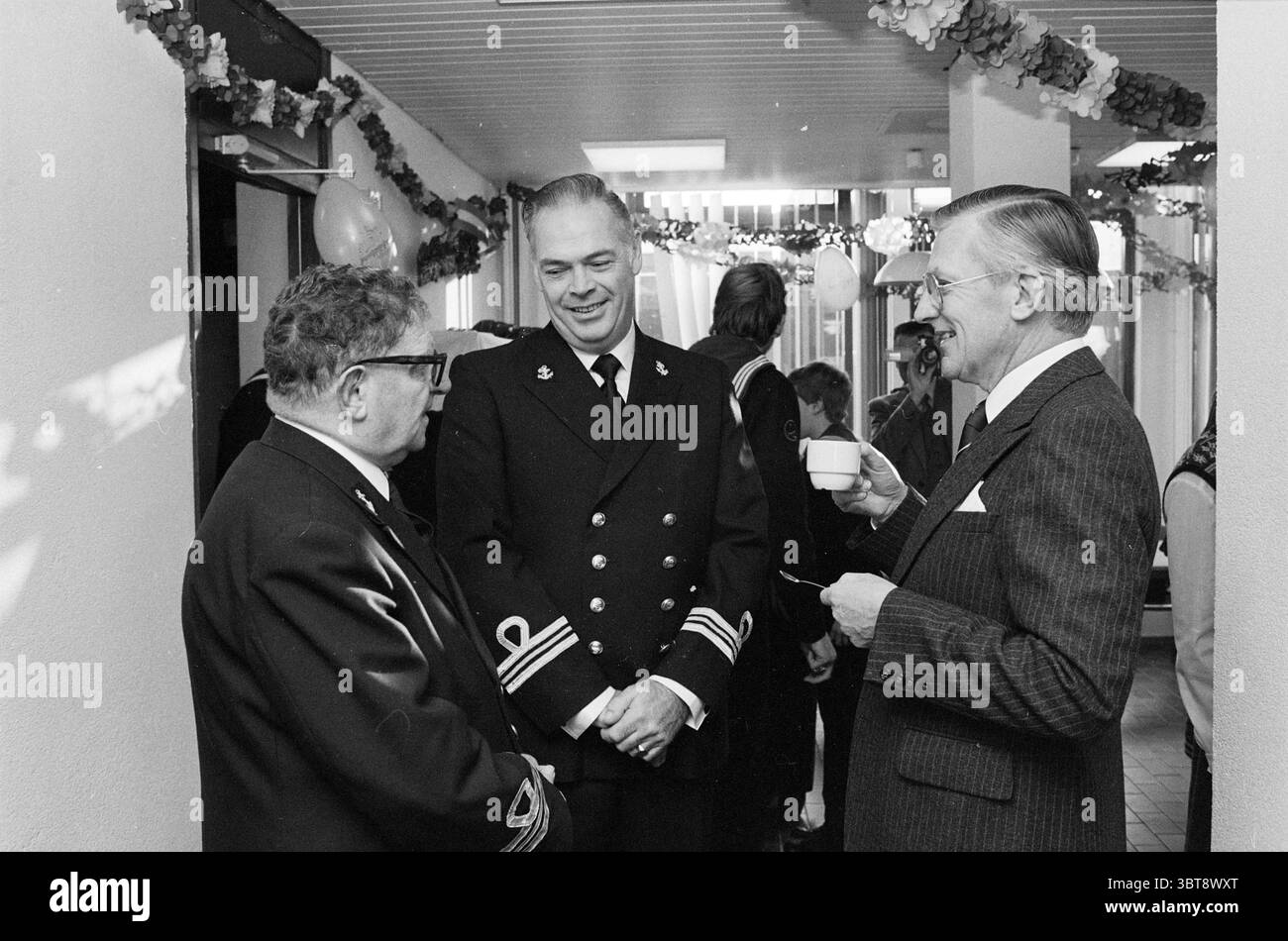 Abschied Arie van de Veer Sea Cadets IJmuiden Sea Scouts IJmuiden the Netherlands, Whizgle News, Dutch Desk, Niederlande, 1950 - 2000 am 12.12.1981. Diese Themen sind in der Abbildung dargestellt. Die Szene zeigt ein Treffen in einem geräumigen Innenraum, möglicherweise in einem Gemeindesaal oder einem ähnlichen Ort, geschmückt mit festlichen Dekorationen. Farbenfrohe Luftschlangen hängen von der Decke, während Ballons verstreut sind, was der Kulisse eine feierliche Atmosphäre verleiht. Drei Männer führen Gespräche an vorderster Front. Eine, eine Sportbrille, trägt einen formellen Anzug mit offenem Kragen, der einen Gelehrten ausstrahlt Stockfoto