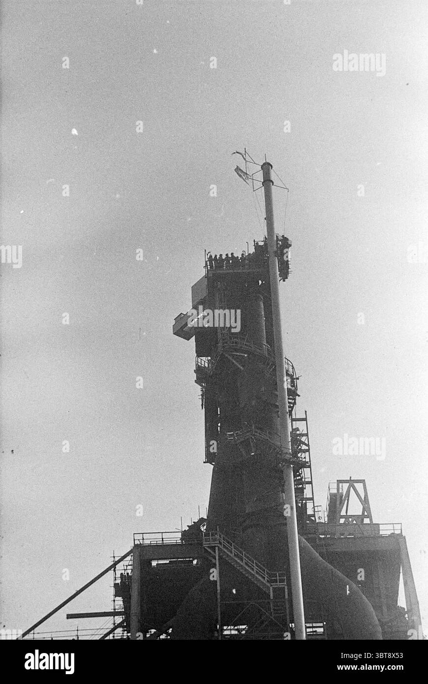 Highlight Blastoven 7 Blastovens, Whizgle News, Dutch Desk, Niederlande, 1950 - 2000 am 03-03-1972. Dies sind die Themen im Bild. Die Szene präsentiert eine hohe Industriestruktur, die sich durch ihre robuste, dunkle Silhouette vor einem klaren, hellen Himmel auszeichnet. Die Struktur verfügt über eine Mischung aus eckigen und gekrümmten Elementen, die die komplexe Konstruktion demonstrieren. Es sind verschiedene Ebenen und Plattformen zu sehen, darunter eine markante Aussichtsplattform oben, auf der zahlreiche Figuren stehen. Diese Zahlen erscheinen klein im Vergleich zu der aufragenden Struktur, was auf ihre immense Größe hindeutet Stockfoto