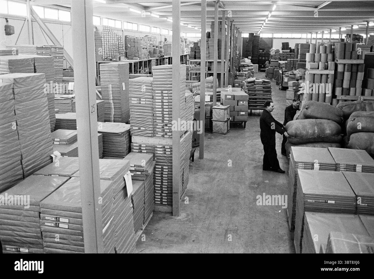 FA.Ruys Verpackungsmaterial kleine Houtweg Haarlem Interiors neue und alte Fabriken Produktionswerkstätten Haarlem kleine Houtweg Niederlande, Whizgle News, Dutch Desk, Niederlande, 1950 - 2000 am 20-12-1962. Diese Themen werden in der Abbildung angezeigt. Die Szene zeigt ein geräumiges Lager, das sich durch eine hohe Decke und gut strukturierte Regalreihen auszeichnet. Die Atmosphäre ist von einem Gefühl von Fleiß erfüllt, der durch die organisierten Stapel von ordentlich verpackten Waren hervorgehoben wird. Die Farbpalette wird von monochromatischen Schattierungen dominiert, vor allem von unterschiedlichen Grautönen, die ein düsteres, aber dennoch f erzeugen Stockfoto