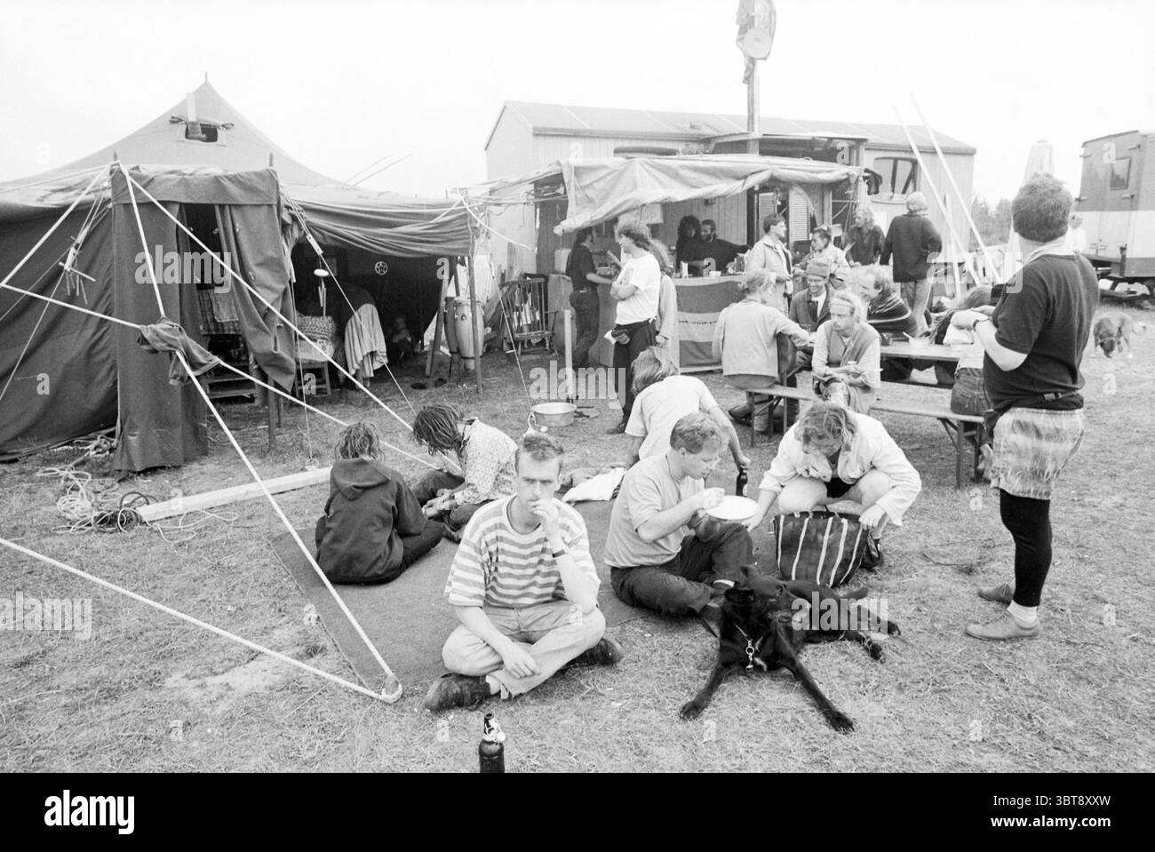 Landjuweel Ruigoord Ruigoord, Whizgle News, Dutch Desk, Niederlande, 1950 - 2000 auf 31-07-1993. Dies sind die Themen im Bild. Die Szene entfaltet sich in einer belebten Umgebung im Freien, die durch eine Mischung aus temporären Strukturen und Einzelpersonen gekennzeichnet ist, die verschiedene Aktivitäten ausüben. Im Hintergrund dienen mehrere Zelte und provisorische Vordächer als Wohn- oder Gemeinschaftsräume. Die Strukturen sind hauptsächlich dunkel und in gedämpfter Farbe gehalten und stehen im Kontrast zum helleren, bewölkten Himmel, der die Szene umgibt. Gruppen von Personen sind in der gesamten Gegend verteilt, einige sitzen auf dem Boden, andere stehen oder bewegen sich Stockfoto