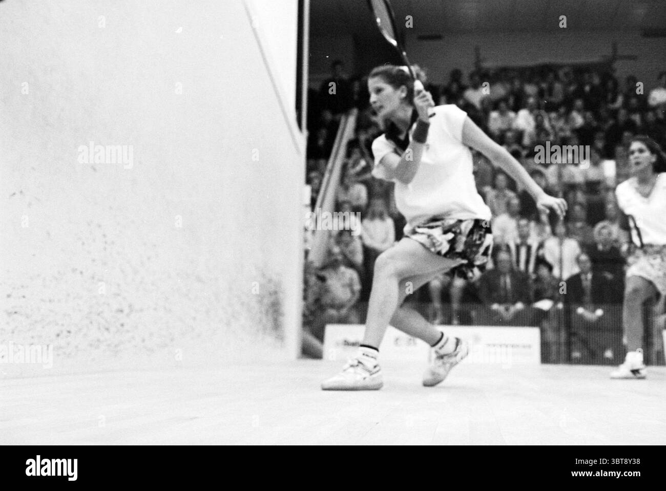 Frauen-Squash-Finale Rulewave [mehr Open], Whizgle News, Dutch Desk, Niederlande, 1950 - 2000 am 23-10-1993. Diese Themen werden in der Abbildung angezeigt. Die Szene fängt einen intensiven Moment in einem Squash-Match ein. Im Vordergrund steht eine Spielerin, die sich in Aktion setzt und ihre Konzentration und Entschlossenheit demonstriert, während sie ihren Schläger schwingt. Ihr Outfit besteht aus einem hellen Oberteil und einem gemusterten Rock, der ein Gefühl von Lebendigkeit in der Wettbewerbsatmosphäre vermittelt. Der Boden ist aus hellem Holz und bietet einen starken Kontrast zu den strukturierten Wänden, die Schrammen und Markierungen von früheren Streichhölzern tragen, was dazu beiträgt Stockfoto