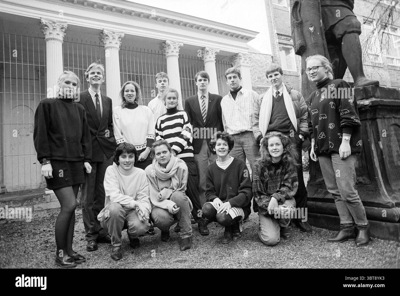Gruppe von Studenten Stadt. Gymnasium Studenten, Whizgle News, Dutch Desk, Niederlande, 1950 - 2000 am 11-01-1988. Das Bild enthält diese Themen. In einer bezaubernden Umgebung im Freien versammelt sich eine Gruppe von 13 Personen eng und schafft eine lebendige Komposition. Der Hintergrund zeigt eine architektonische Struktur mit hohen, weißen Säulen, die an einen klassischen Stil erinnern, eingerahmt von Ziegelwänden, die Textur verleihen. Die allgemeine Atmosphäre ist lässig und entspannt. Die Individuen sind in einer Mischung aus lässiger und halbformaler Kleidung gekleidet, wobei die Kleidungsstile eine Vielzahl von Mustern und Farben widerspiegeln. Also Stockfoto