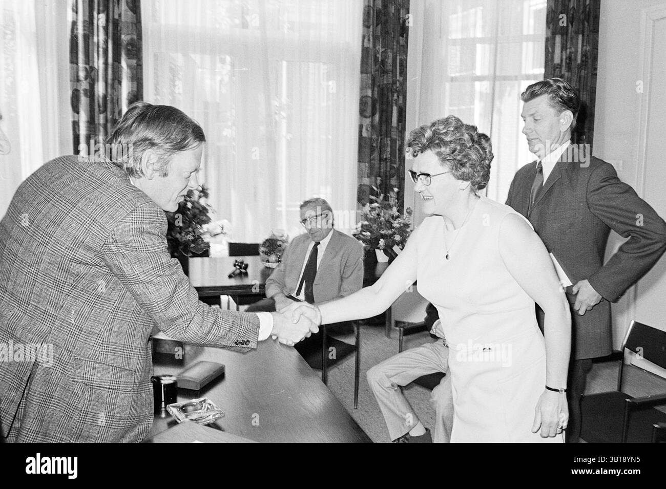 Drei Schullehrer zu ehren. Ehren, Whizgle News, Dutch Desk, Niederlande, 1950 - 2000 am 23-06-1972. Das Bild enthält diese Themen. Die Szene verfügt über eine formelle Inneneinrichtung, die durch einen großen, gut beleuchteten Raum gekennzeichnet ist. Sonnenlicht filtert durch große Fenster, wirft weiche Schatten auf den Boden und verbessert die Helligkeit insgesamt. Im Vordergrund stehen sich zwei Personen in einem engagierten Handschlag zueinander gegenüber. Eine Person, die in einem strukturierten Karomuster gekleidet ist, scheint mittleren Alters zu sein und eine Brille trägt. Die andere ist eine Frau in einem ärmellosen, hellen Kleid mit einer selbstbewussten Haltung Stockfoto