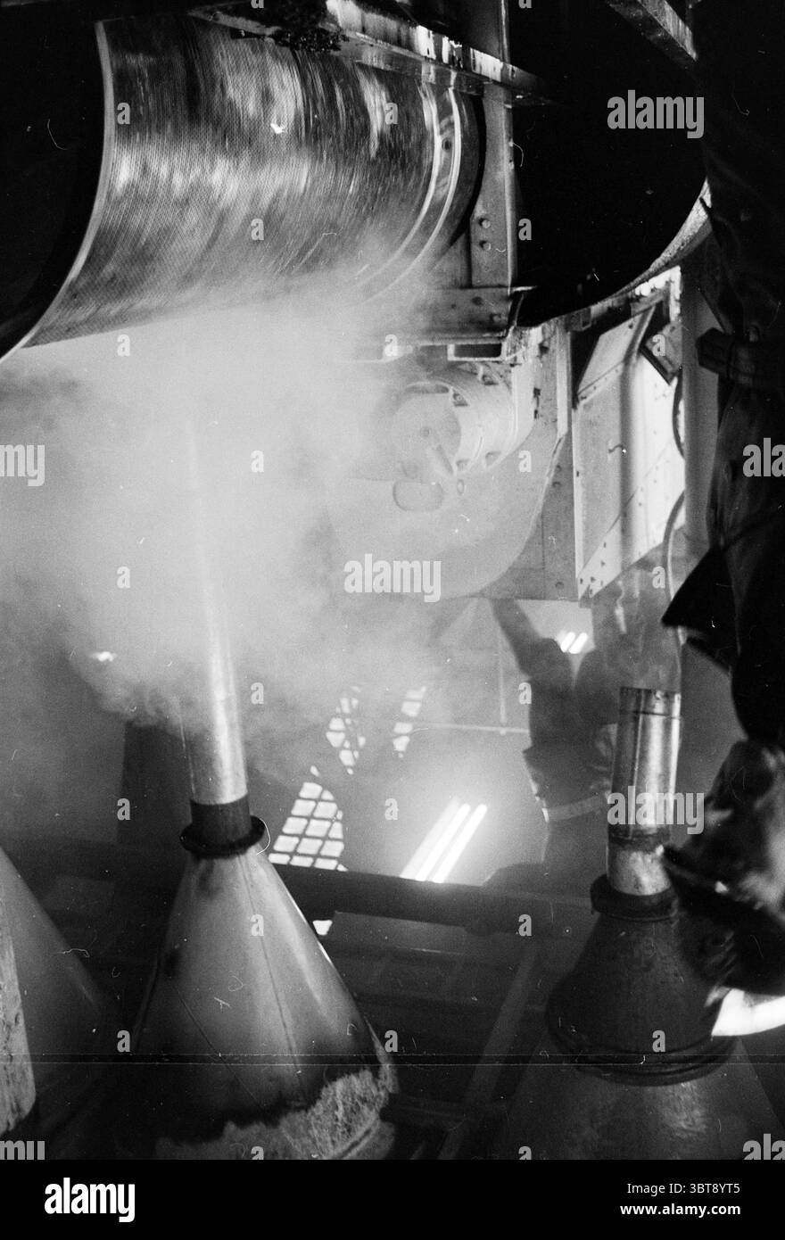 Feuer Kakaobrenner in der Feuerfeuerwehr, Whizgle News, Dutch Desk, Niederlande, 1950 - 2000 am 08-01-1963. Dies sind die Themen im Bild. Die Szene zeichnet sich durch ein Gefühl von Fleiß und Komplexität aus, das in einer monochromatischen Palette festgehalten wird, die den Kontrast zwischen Licht und Schatten betont. Die Umgebung ist mit großen, komplizierten Maschinen gefüllt, die hauptsächlich aus Metall und Stahl bestehen. Im Vordergrund sind Teile von Rohren und Trichtern zu sehen, von denen einige sich nach unten verjüngen, während andere sich verbiegen und verdrehen, was auf ein Netzwerk von Arbeitsabläufen hindeutet. Die Flächen haben ein s Stockfoto