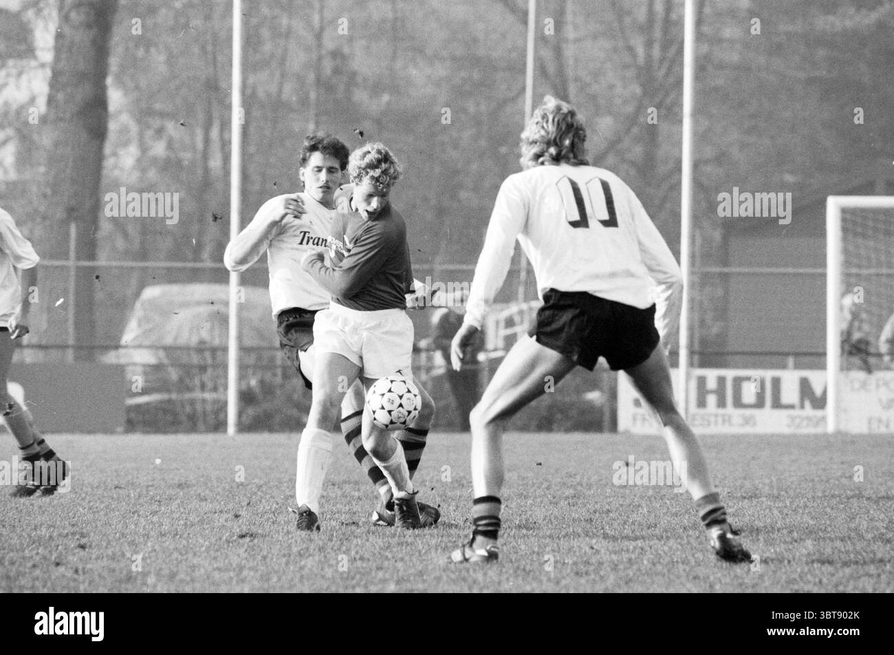 RCH - Stormvogels Football R.C.H., Whizgle News, Dutch Desk, Niederlande, 1950 - 2000 am 29.11.1986. Das Bild enthält diese Themen. Die Szene fängt einen dynamischen Moment in einem Fußballspiel auf einem grasbewachsenen Feld ein, das im Hintergrund leicht unscharf ist und auf Bewegung und Aktivität hinweist. Ein Gefühl der Dringlichkeit wird vermittelt, wenn drei Spieler ein angespanntes Spiel führen. Im Vordergrund befinden sich zwei Spieler in unmittelbarer Nähe. Einer, der in einem auffälligen roten Trikot gekleidet ist, versucht, die Kontrolle über den dazwischen liegenden Fußball zu gewinnen. Der andere Spieler in einem weißen Trikot mit dem Wort "Trans Stockfoto