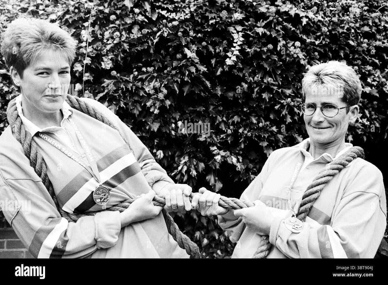 TUG-of-war wird Champion Santpoort Santpoort, Whizgle News, Dutch Desk, Niederlande, 1950 - 2000 am 02.10.1997. Das Bild enthält diese Themen. In dieser Szene stehen zwei Individuen nebeneinander, jeder packt gegenüberliegende Enden eines dicken, schweren Seils, was auf ein verspieltes Tauziehen hindeutet. Sie teilen einen leichtsinnigen Ausdruck; einer hat ein Lächeln, während der andere entschlossen und doch amüsant erscheint. Beide tragen passende sportliche Oberteile mit breiten Streifen in leuchtenden Farben, die das Gefühl von Kameradschaft und Wettkampf verstärken. Der Hintergrund ist mit üppigem Grün gefüllt, mit einer Hecke dicht mit Blättern, p Stockfoto