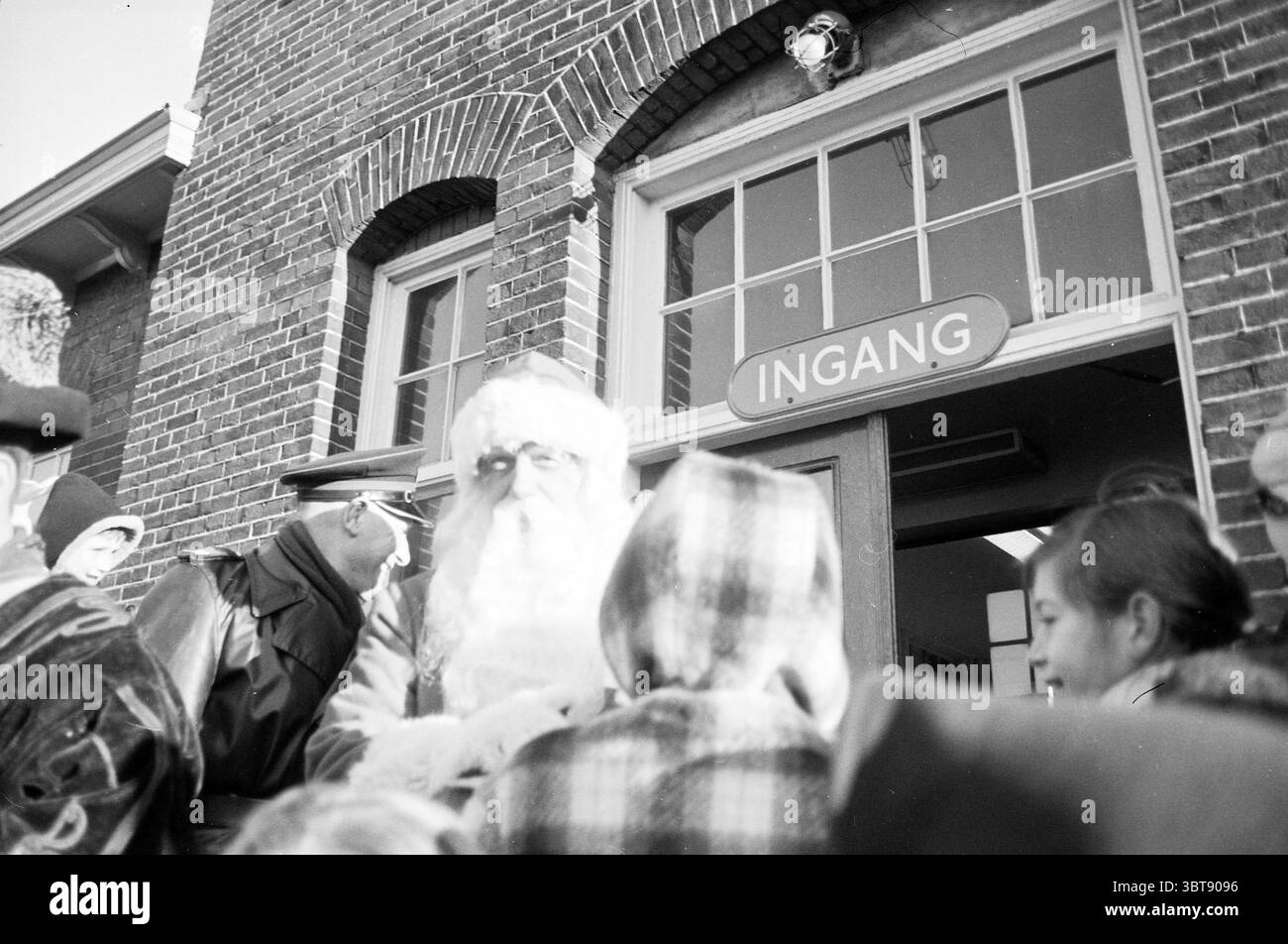 Weihnachtsmann vor dem Bahnhof Bloemendaal Weihnachten Weihnachtsdekoration Weihnachtsdekoration Weihnachtsdekoration, Whizgle News, Dutch Desk, Niederlande, 1950 - 2000 am 27-12-1962. Diese Themen sind in der Abbildung dargestellt. Die Szene fängt ein lebhaftes Treffen am Eingang eines Gebäudes ein, das durch seine rote Backsteinfassade gekennzeichnet ist. Die Architektur verfügt über große, bogenförmige Fenster, die natürliches Licht hereinlassen und ein ruhiges Leuchten auf die Gesichter der umliegenden Personen werfen. Eine festliche Atmosphäre ist offensichtlich, verstärkt durch die Anwesenheit einer Figur, die als Weihnachtsmann verkleidet ist, der mit seinem langen weißen Bären große Aufmerksamkeit auf sich zieht Stockfoto