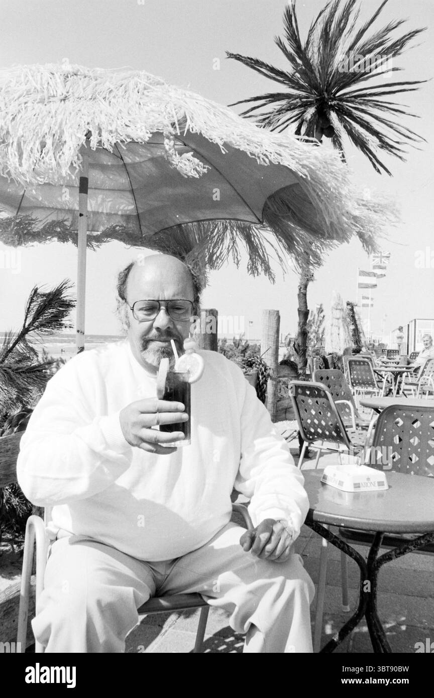 Simon van Collem with a Drink Personen t.v., Whizgle News, Dutch Desk, Niederlande, 1950 - 2000 am 18-06-1983. Die Abbildung zeigt diese Themen. Die Szene fängt eine sonnendurchflutete Umgebung im Freien ein, wahrscheinlich am Strand oder an einer belebten Promenade. Im Vordergrund sitzt ein Mann mittleren Alters mit rundem Gesicht und Brille an einem kleinen runden Tisch. Er trägt ein helles Sweatshirt, das eine entspannte Atmosphäre vermittelt. Der Mann wiegt ein hohes Glas, gefüllt mit einem dunklen Getränk, geschmückt mit einer Scheibe Zitrone und möglicherweise einem Strohhalm, was ein erfrischendes Getränk an einem warmen Tag andeutet. Er umgibt lebendige Elemente des Sommers. A Large Stockfoto
