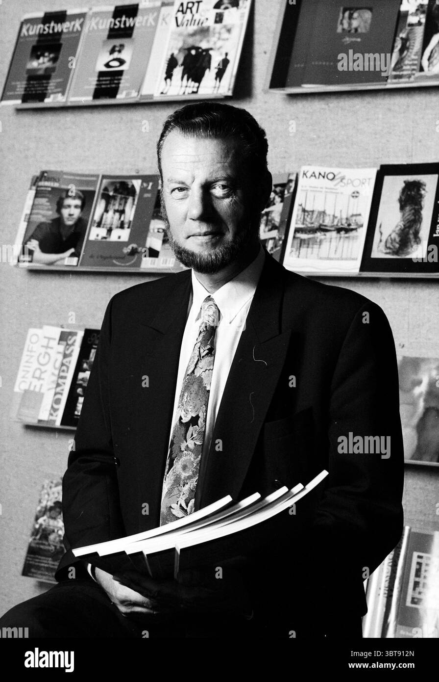 Mr. Mast File Letter, Whizgle News, Dutch Desk, Niederlande, 1950 - 2000 am 25-08-1996. Die Abbildung zeigt diese Themen. In dieser Szene steht ein Mann selbstbewusst in einem formellen schwarzen Anzug mit einer charakteristischen Krawatte mit einem bunten Muster. Sein Ausdruck ist zielstrebig und spiegelt ein Gefühl von Professionalität und Ernsthaftigkeit wider. Der Hintergrund besteht aus einer Wand mit einer Reihe von Zeitschriften und Kunstpublikationen, die eine Vielzahl von Covern in gedämpften Tönen zeigen, hauptsächlich Graustufen mit gelegentlichen Farbakzenten. Dies schafft einen interessanten Kontrast zu der Kleidung des Mannes, sowie zu der sub d Stockfoto