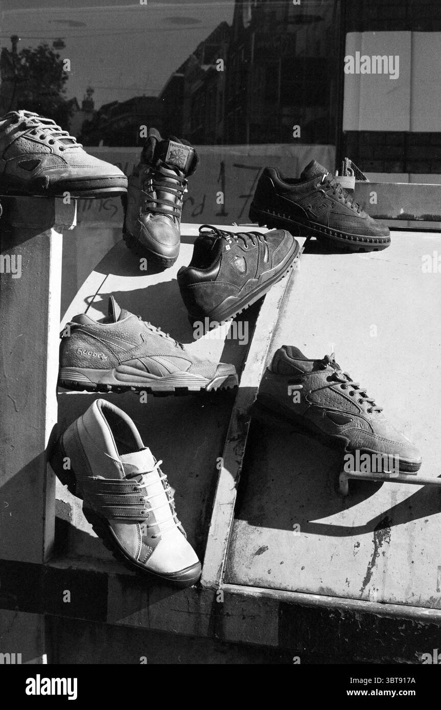 Schuhe von Foot locker Store, Whizgle News, Dutch Desk, Niederlande, 1950 - 2000 am 08-08-1991. Diese Themen werden in der Abbildung angezeigt. Die Szene zeigt eine Kollektion von Schuhen, die in einer etwas willkürlichen Art und Weise angeordnet sind, wodurch eine faszinierende visuelle Komposition entsteht. Sie liegen vor einem Hintergrund, der industriell wirkt, wahrscheinlich einer Wand oder einer Struktur mit strukturierter Oberfläche. Die Schuhe variieren stark im Stil, einschließlich sportlicher Turnschuhe, formeller Schuhe und lässiger Designs. Jedes Paar zeigt unterschiedliche Farben, hauptsächlich in Schwarz-, Braun- und Grautönen, mit kontrastierenden Texturen wie Leder und Stockfoto