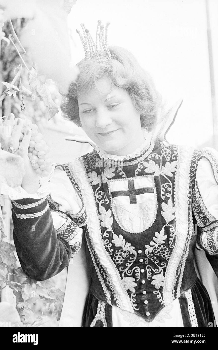 Wine Festival Unkel Germany Wine, Whizgle News, Dutch Desk, Niederlande, 1950 - 2000 am 09-10-1977. Dies sind die Themen im Bild. Die Szene zeigt eine junge Frau in einem aufwendigen Kostüm, das sich durch komplizierte Details und lebendige Texturen auszeichnet. Ihre Kleidung besteht aus einem dunklen Samtkleid, das mit verzierten Blumenmustern verziert ist und einen eleganten Kontrast zu helleren Elementen bildet. Eine königliche Krone sitzt auf ihrem welligen Haar und verleiht ihrem Aussehen einen Hauch von Adel. In ihrer rechten Hand hält sie zart einen Traubenstrauch, deren abgerundete Formen und üppige Texturen ein Gefühl vermitteln Stockfoto