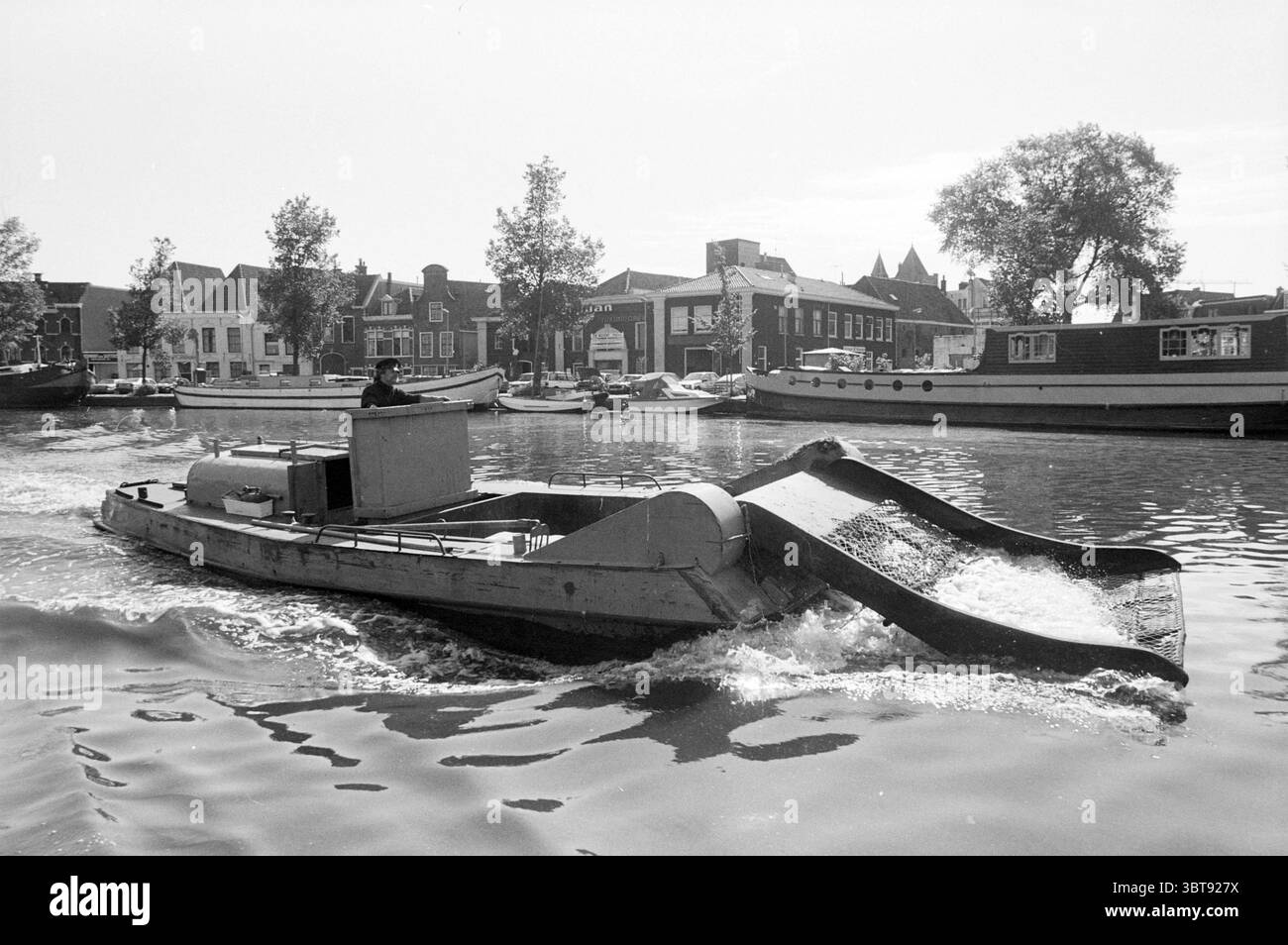 Bootstour mit Hafenmeister durch den Spaarne Kanal Kreuzfahrt Besichtigungstouren Sightseeing Flüge Haarlem Niederlande, Whizgle News, Dutch Desk, Niederlande, 1950 - 2000 am 01-07-1987. Dies sind die Themen im Bild. In der Szene gleitet ein kleines Boot über ein glitzerndes Wasser und erzeugt sanfte Wellen, die das Sonnenlicht reflektieren. Das Boot ist mit einem flachen, offenen Deck und einer besonderen Form ausgestattet, mit einem breiten Bug, der durch die Wellen zu drücken scheint. Am Steuer steht eine Figur, die sich vielleicht auf die Navigation konzentriert, in lässiger Kleidung, die für einen Tag auf dem Wasser geeignet ist. Umgebung Stockfoto