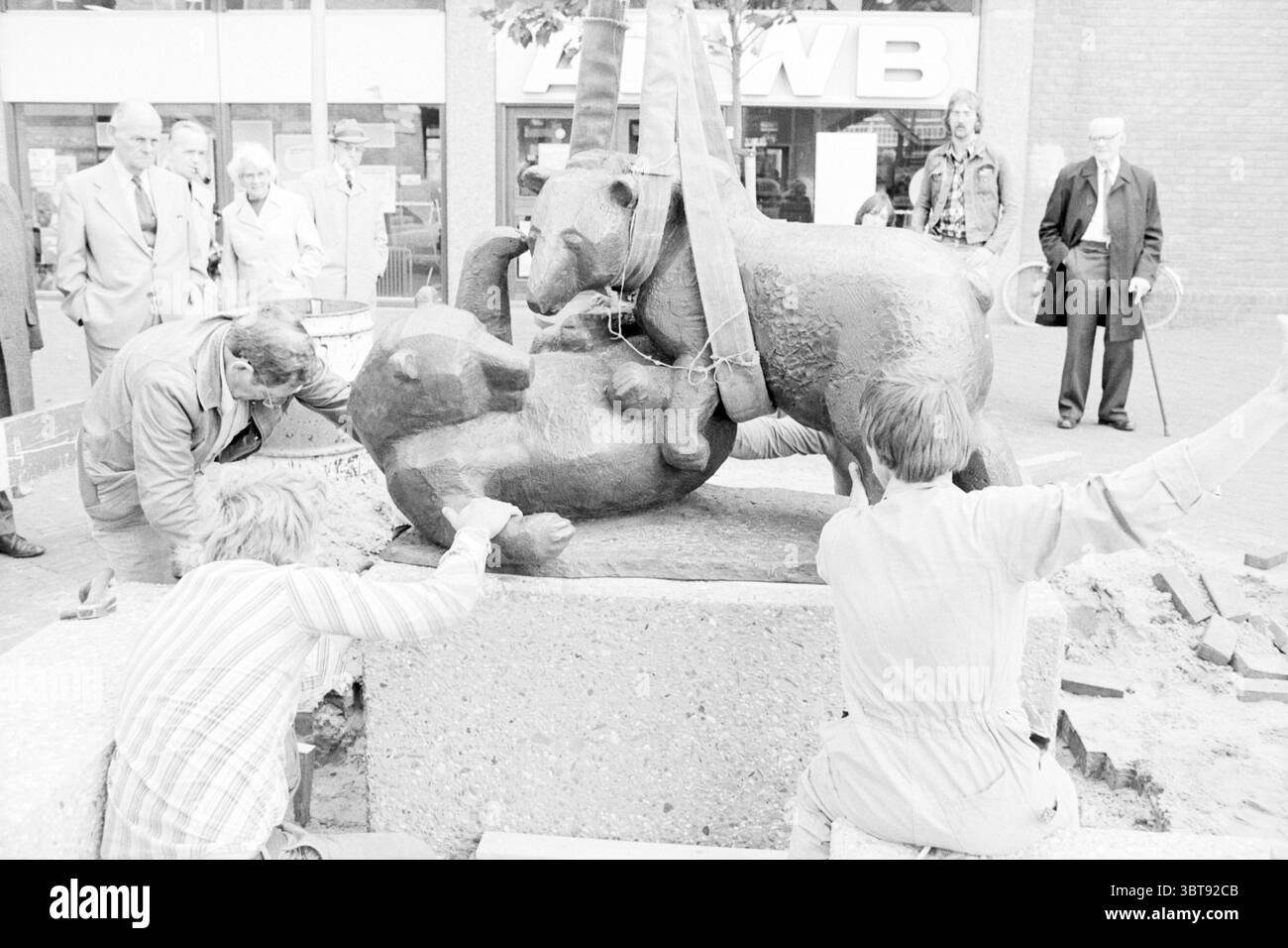 Place image Bears Haarlem Stationsplein the Netherlands, Whizgle News, Dutch Desk, Niederlande, 1950 - 2000 am 04-09-1978. Diese Themen werden in der Abbildung angezeigt. Die Szene zeigt eine geschäftige urbane Umgebung, die sich durch eine Mischung aus Aktivität und Fokus auszeichnet. Im Vordergrund hocken sich zwei Individuen neben einer großen Skulptur, die zwei stilisierte Tiere zeigt, möglicherweise Hunde, die spielerisch miteinander interagieren. Sie sind in Arbeitskleidung gekleidet, was darauf hindeutet, dass sie an der Installation oder Wartung der Skulptur beteiligt sind. Ihre konzentrierten Ausdrucksformen deuten auf eine Hingabe an die anstehende Aufgabe hin. S Stockfoto