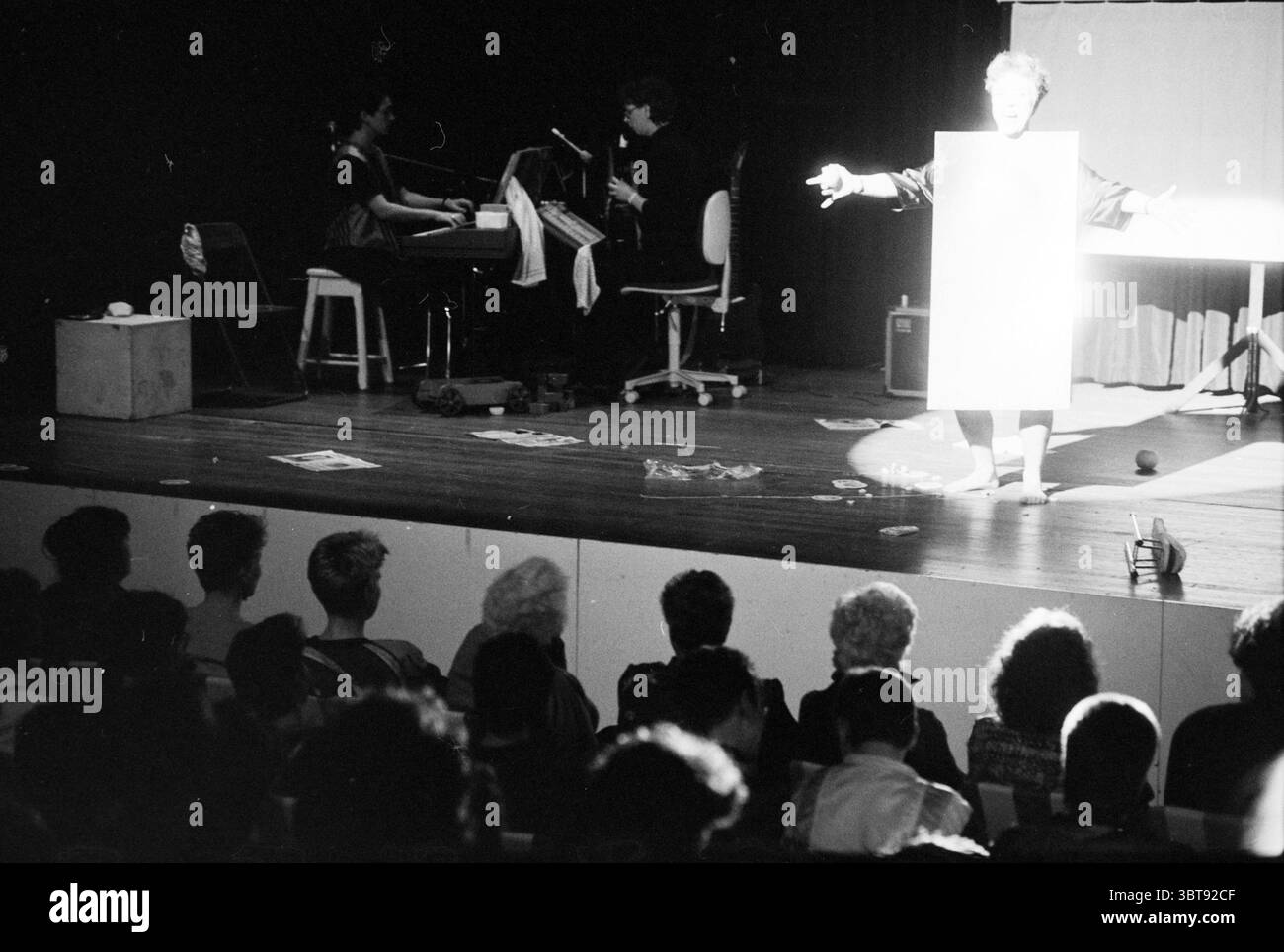 Women's Day Concert Hall Women, Whizgle News, Dutch Desk, Niederlande, 1950 - 2000 am 08-03-1990. Dies sind die Themen im Bild. Die Szene fängt eine Theateraufführung auf einer schwach beleuchteten Bühne ein. In der Mitte steht ein Darsteller mit ausgestreckten Armen, gekleidet in einem knallweißen Outfit, das einen scharfen Kontrast zur umgebenden Dunkelheit bildet. Das helle Licht von oben beleuchtet ihre Figur und schafft einen markanten Fokus inmitten der schattigen Kulisse. Links ist ein kleines Ensemble zu sehen, mit einem Pianisten auf einem Keyboard und einem anderen Musiker in der Nähe Stockfoto