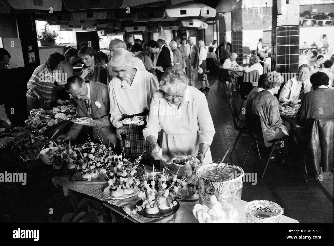 Patient Market Provincial Government Building Emergency Service Assistance Shelter etc. Market, Whizgle News, Dutch Desk, Niederlande, 1950 - 2000 auf 28-09-1986. Diese Themen werden in der Abbildung angezeigt. Die Szene fängt ein lebhaftes Treffen in einem geräumigen Speisesaal mit Tischen, die mit einer Auswahl an bunten Gerichten geschmückt sind, ein. Gäste jeden Alters mischen sich, ihr Ausdruck vermittelt Freude und Begeisterung. Hell dekorierte Essensplatten, darunter lebendige Früchte und sorgfältig angeordnete Vorspeisen, stehen im Mittelpunkt der Komposition. Eine große Schüssel steht gut sichtbar, gefüllt mit einem bunten Salat, während Stockfoto