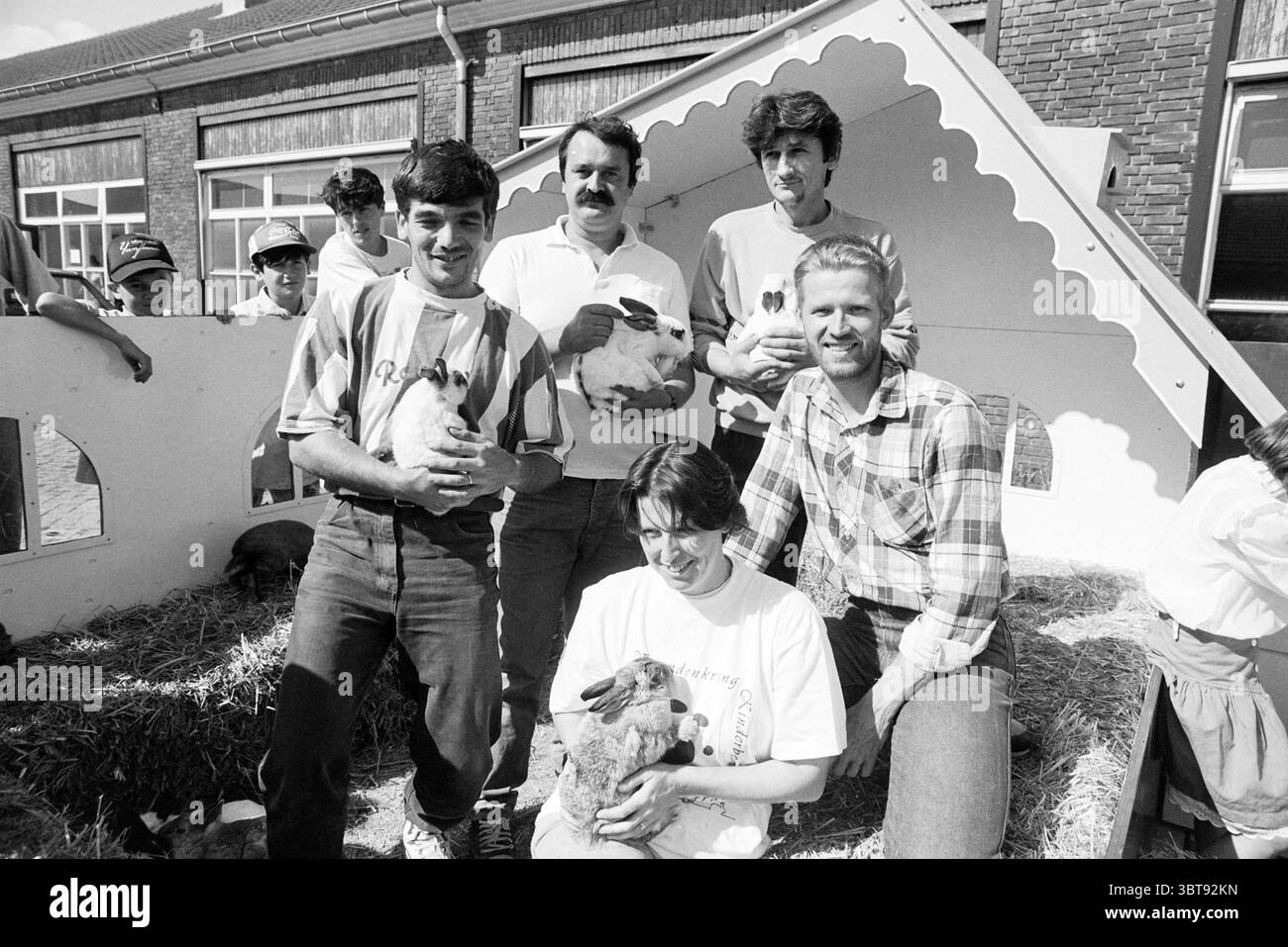 Streichelzoo Bosnians, Whizgle News, Dutch Desk, Niederlande, 1950 - 2000 am 16-08-1993. Das Bild enthält diese Themen. Die Szene zeigt eine Gruppe von sechs Individuen, die eine angenehme Interaktion mit Kaninchen führen. Die Lage scheint ein Außenbereich mit rustikalem Charme zu sein, möglicherweise in der Nähe einer Scheune oder einer ähnlichen Struktur, gekennzeichnet durch robuste Backsteinmauern und ein schräges Dach. Im Vordergrund stehen vier Männer, die jeweils ein Kaninchen halten und ihre Verbindung zu den Tieren zeigen. Die Einzelpersonen tragen legere Kleidung, die eine entspannte Atmosphäre widerspiegelt; einige sind in L Stockfoto