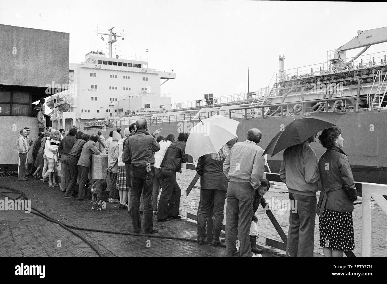 Schlechtes Wetter IJmuiden Wetter Wetter Service IJmuiden Velsen IJmuiden Niederlande, Whizgle News, Dutch Desk, Niederlande, 1950 - 2000 am 21-07-1981. Das Bild enthält diese Themen. Eine Gruppe von Einzelpersonen steht neben einem Dock und blickt alle aufmerksam auf ein großes Schiff, das in der Nähe vertäut ist. Die Gruppe, die sich aus Männern und Frauen zusammensetzt, zeigt eine Reihe von Ausdrucksformen, von Neugier bis hin zur Begeisterung. Verschiedene Regenschirme, einige geschlossen und andere offen, bieten einen Spritzer weißer und heller Töne vor dem Hintergrund grauer Himmel, was auf einen drohenden oder anhaltenden Nieselregen hindeutet. Die Leute sind in einer Mixtur gekleidet Stockfoto
