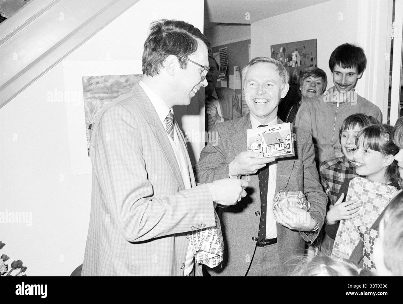 Inbetriebnahme der Wohnhalle Haarlem Housing Commissioning Inbetriebnahme Haarlem the Netherlands, Whizgle News, Dutch Desk, Niederlande, 1950 - 2000 am 14-03-1984. Dies sind die Themen im Bild. Die Szene zeigt ein lebhaftes Indoor-Treffen, wahrscheinlich eine Feier wie ein Geburtstag oder Feiertag. Zwei Erwachsene Männer stehen im Zentrum, beide lächeln breit und deuten auf einen Moment der Freude oder des Lachens hin. Ein Mann, gekleidet in einem hellen Anzug mit Karomuster, scheint ein Geschenk zu überreichen, während der andere in einem dunkleren Anzug ein Paket hochhält. Sein Ausdruck strahlt Erregung aus Stockfoto