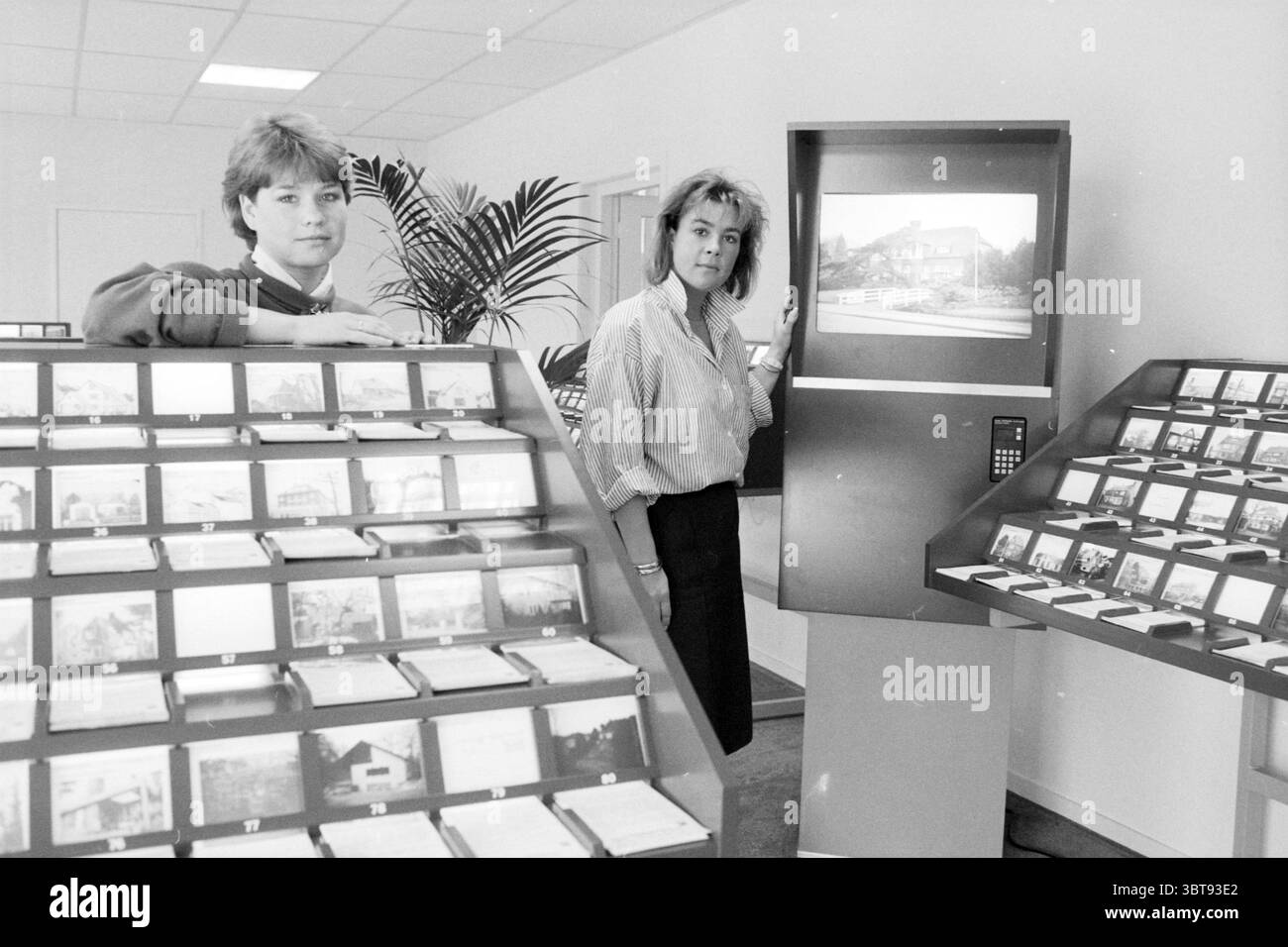 Digitales Angebot von Häusern zum Verkauf, Whizgle News, Dutch Desk, Niederlande, 1950 - 2000 auf 05-1988. Diese Themen sind in der Abbildung dargestellt. In einem gut beleuchteten Raum stehen zwei Frauen vor einer Ausstellung organisierter Karten, was auf eine Umgebung im Zusammenhang mit Immobilien oder Tourismus hindeutet. Die Frauen befinden sich auf beiden Seiten eines digitalen Bildschirms, auf dem ein Foto, möglicherweise eines Grundstücks oder einer Landschaft, dargestellt wird, was auf visuelle Informationen hinweist. Die Frau auf der linken Seite hat kurze, gestylte Haare und trägt ein dunkles Oberteil mit Kragen, das ein Gefühl von Professionalität ausstrahlt. Ihr Ausdruck ist neutral, da sie direkt auf t schaut Stockfoto