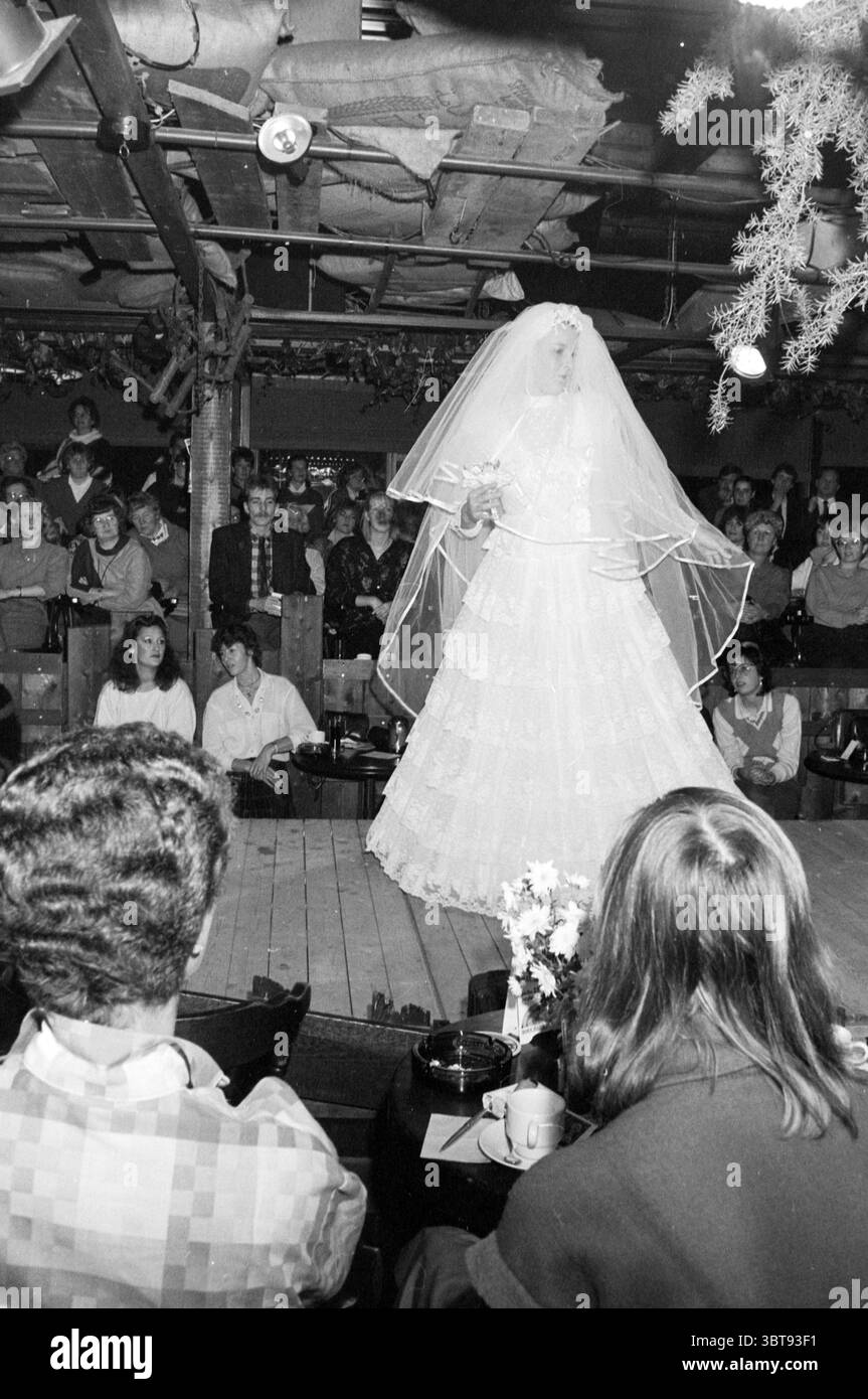 Fashion Show Brautmode im Claus Party House Haarlem Orionweg the Netherlands, Whizgle News, Dutch Desk, Niederlande, 1950 - 2000 am 25-11-1985. Dies sind die Themen im Bild. In einem schwach beleuchteten Veranstaltungsort posiert ein Brautmodell anmutig auf einer Holzbühne in einem raffinierten weißen Hochzeitskleid, das in Schichten kaskadiert. Das Kleid ist zart, mit komplizierten Spitzendetails und einem geschwungenen Zug, der ihrer Silhouette Eleganz verleiht. Ein langer Schleier fließt über ihren Rücken, umrahmt ihr Gesicht und unterstreicht die Mystik des Brautlooks. Die Szene ist voller Vorfreude, während ein Publikum sitzt Stockfoto