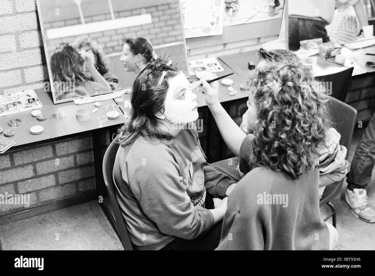 Face Painting in Progress, Whizgle News, Dutch Desk, Niederlande, 1950 - 2000 im Jahr 1990. Die Abbildung zeigt diese Themen. In einem lebendigen Innenbereich mit freiliegenden Ziegelwänden werden zwei Personen in einem Moment der Verwandlung gefangen genommen. Der Raum wirkt lebendig und doch lässig, mit hellen Postern und verschiedenen Objekten, die über Tische verteilt sind, verziert, was auf eine kreative Atmosphäre hindeutet. Eine Person sitzt auf einem Stuhl, ihr Blick ist aufmerksam, während die andere sich schminkt. Die Sitzperson hat eine entspannte Haltung und ist in einem gemütlichen, übergroßen Kleidungsstück mit weichen Farbtönen dekoriert, die den dunklen Ton abheben Stockfoto