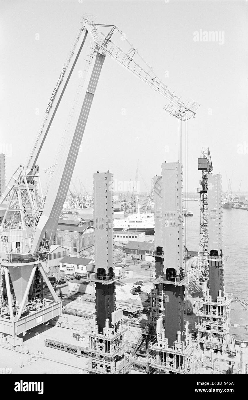 Crane Island Crane in Schiedam Crane Islands, Whizgle News, Dutch Desk, Niederlande, 1950 - 2000 am 03-09-1964. Diese Themen sind in der Abbildung dargestellt. Die Szene wird von einer Industrielandschaft dominiert, die von hochaufragenden Kränen und großen, imposanten Bauwerken geprägt ist. Die Kraniche mit ihren langen Gelenkarmen reichen hoch in den Himmel und werfen lange Schatten auf den Boden. Sie sind in einem schlichten weiß lackiert und stehen in starkem Kontrast zu den dunklen Oberflächen der Maschinen, die sie überwachen. Im Vordergrund stehen mehrere vertikale Stahlsäulen hoch und glitzern unter dem hellen Licht. Th Stockfoto