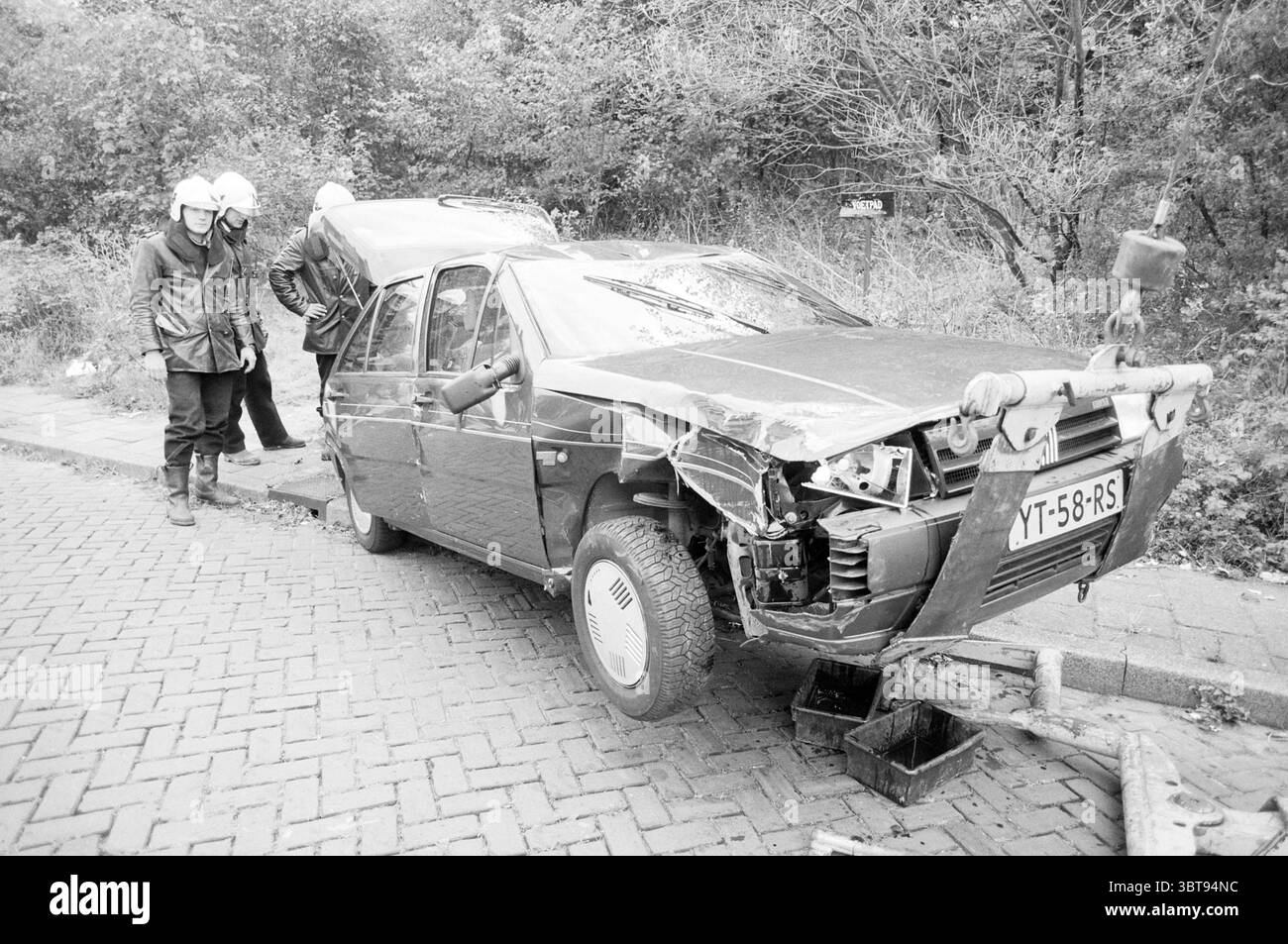 Autounfall in der Maasstraat IJmuiden Maasstraat Niederlande, Whizgle News, Dutch Desk, Niederlande, 1950 - 2000 am 06-10-1991. Die Abbildung zeigt diese Themen. Die Szene zeigt ein beschädigtes Auto, das neben einer Kopfsteinpflasterstraße steht, die leicht mit Gras und Pflanzen bewachsen ist. Das Fahrzeug hat erhebliche Schäden an der Front erlitten, wobei die zerknitterte Motorhaube und die zersplitterte Windschutzscheibe auf einen Zusammenstoß hindeuten. Das dunkle Äußere wird durch die stumpfen, gedämpften Töne der Umgebung kontrastiert, die die düstere Stimmung verstärken. Im Vordergrund zwei Personen in Schutzkleidung Stockfoto