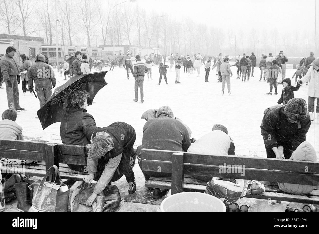 Herzlichen Glückwunsch an den 100000. Besucher der Eisbahn Haarlem Eislaufbahn Eishockey Haarlem Niederlande, Whizgle News, Dutch Desk, Niederlande, 1950 - 2000 am 22-01-1984. Dies sind die Elemente im Bild. Die Szene ist voller Aktivität, spielt auf einer gefrorenen Eisbahn im Freien, umgeben von einem weichen, winterlichen Dunst. Im Hintergrund gleiten die Skater über das Eis, einige bewegen sich anmutig, während andere eher zögerlich wirken. Die Skater tragen eine Mischung aus lebendiger Winterbekleidung – Jacken in Rot, Blau und Gelb – im Kontrast zum schneebedeckten Untergrund. In Th Stockfoto