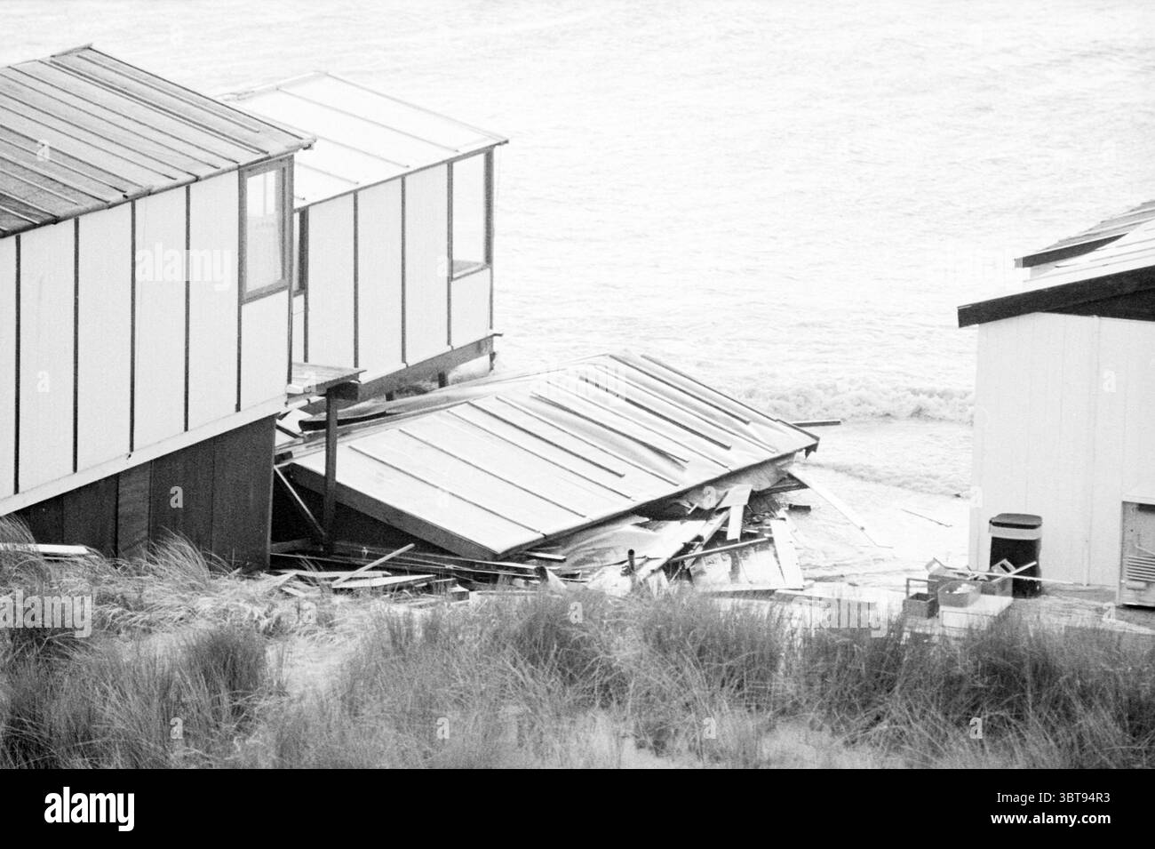 Storm Beach IJmuiden Storm and Storm Damage Strand und Strände IJmuiden Niederlande, Whizgle News, Dutch Desk, Niederlande, 1950 - 2000 am 19-04-1980. Das Bild enthält diese Themen. Die Szene fängt eine Küstenlandschaft ein, die durch eine schlichte, monochrome Palette gekennzeichnet ist, die ein Gefühl der Verzweiflung vermittelt. Eingebettet in eine Sanddüne, steht eine Ansammlung von Küsten in verschiedenen zustand der Verwerfung. Die Hütten weisen schlichte, kastenförmige Formen mit schrägen Dächern auf, deren Außenflächen in verwitterten weiß- und Grautönen gestrichen sind. Im Vordergrund befinden sich verstreute Trümmer, darunter Holzdielen und Teile Stockfoto