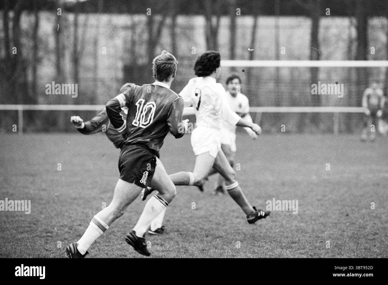 Football Match, Whizgle News, Dutch Desk, Niederlande, 1950 - 2000 am 12-1988. Diese Themen werden in der Abbildung angezeigt. Die Szene fängt einen dynamischen Moment auf einem Fußballfeld ein, der durch eine Mischung aus Bewegung und Fokus gekennzeichnet ist. Im Mittelpunkt der Komposition stehen zwei Spieler in der Mitte der Action. Der eine trägt ein leuchtendes rotes Trikot mit der Nummer 10 und zeigt entschlossene Leichtathletik, während er über das üppige grüne Gras sprintet. Sein Haar ist leicht zerzaust, was auf die Intensität des Spiels hindeutet. Der gegnerische Spieler, der in einem weißen Trikot gekleidet ist, wird weiter positioniert, wobei die Nummer 5 auf dem Rücken sichtbar ist. Sein Stockfoto