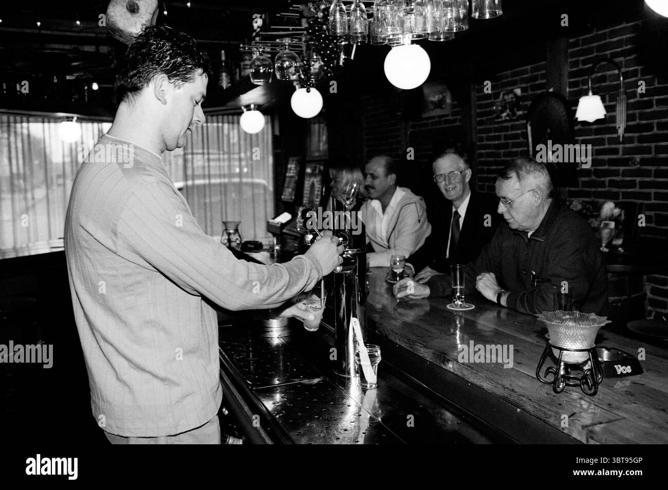 Café de Tapperij, Whizgle News, Dutch Desk, Niederlande, 1950 - 2000 am 27-02-2001. Das Bild enthält diese Themen. In einer gemütlichen, schwach beleuchteten Bar ist die Atmosphäre warm und einladend, die durch satte Holztöne und das sanfte Leuchten der Hängeleuchten vermittelt wird. Ein junger Barkeeper in legeren Kleidern konzentriert sich darauf, ein Getränk aus einem glänzenden Wasserhahn zu gießen. Seine Haltung deutet auf Hingabe an sein Handwerk hin, mit einer leichten Neigung zur Theke, die seine Auseinandersetzung mit der anstehenden Aufgabe unterstreicht. Die Bar selbst ist poliert und reflektierend, mit einer Reihe sauberer Gläser, die ordentlich über der Theke gestapelt sind. Be Stockfoto