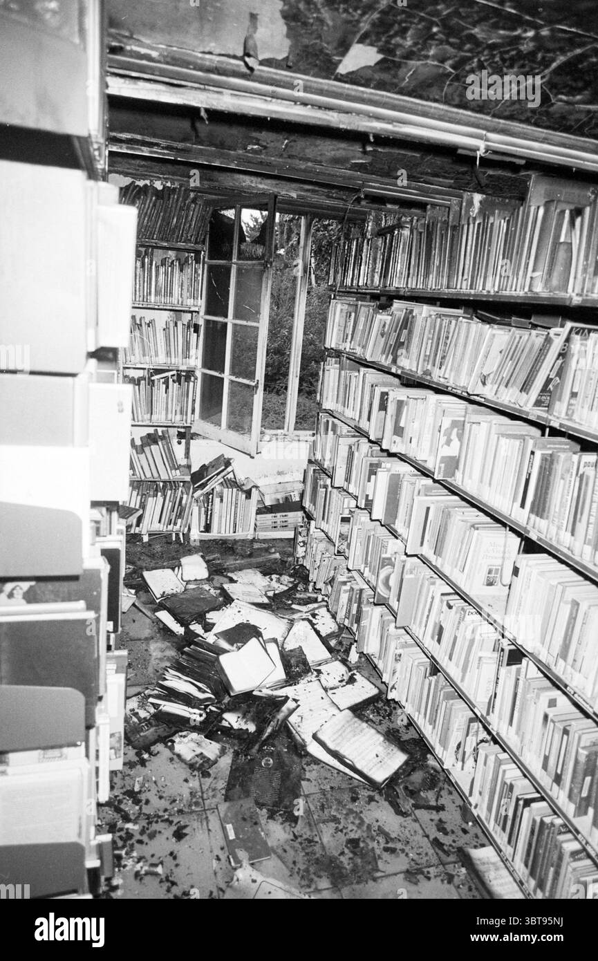 Brandschaden im Haus in Zaanen Feuer Feuerwehr Haarlem Orionweg Niederlande, Whizgle News, Dutch Desk, Niederlande, 1950 - 2000 am 13-10-1989. Das Bild enthält diese Themen. In einem schwach beleuchteten Raum erstrecken sich mit Büchern gesäumte Regale zur Decke, deren Stacheln meist in verblassenen Farben und verschiedenen Größen bedeckt sind. Die Regale bilden einen schmalen Korridor, der das Auge zu einem Fenster am anderen Ende führt, wo weiches natürliches Licht durchdringt und die in der Luft schwebenden Staubhäute beleuchtet. Der Boden ist überfüllt mit verstreuten Papierbögen, einige zerknittert und zerrissen Stockfoto