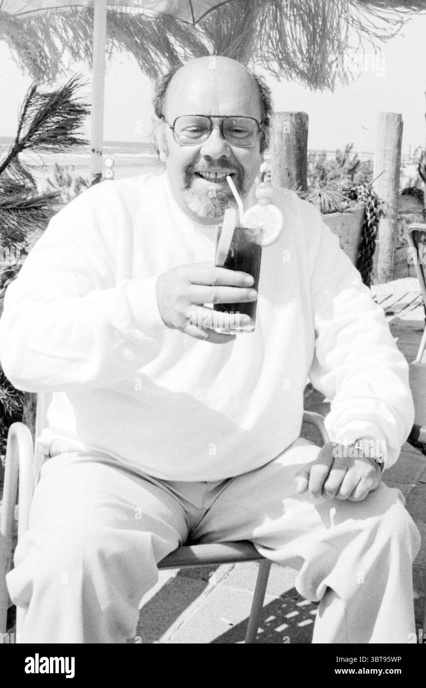 Simon van Collem with a Drink Personen t.v., Whizgle News, Dutch Desk, Niederlande, 1950 - 2000 am 18-06-1983. Das Bild enthält diese Themen. In einer sonnigen Umgebung im Freien sitzt ein fröhlicher Mann bequem in einem zwanglosen Stuhl und strahlt Wärme und Genuss aus. Er hat ein rundes Gesicht, eingerahmt von dünnen Brillen, die leicht im Licht schimmern. Sein Ausdruck ist hell und zeigt ein breites Lächeln, während er ein Glas mit einem dunklen Getränk hochhält, das mit einer Scheibe Zitrone und einem Strohhalm geschmückt ist, was auf ein erfrischendes Getränk hindeutet. Der Mann trägt einen kuscheligen weißen Pullover, der einen Kontrast zu den weichen, sandigen Farbtönen bildet Stockfoto