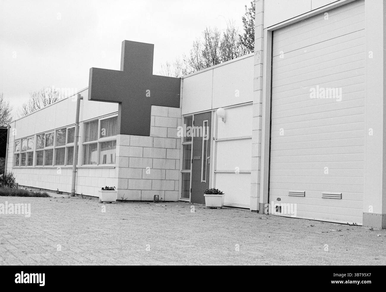 Red Cross Building Velsen Velsen, Whizgle News, Dutch Desk, Niederlande, 1950 - 2000 am 03-05-1994. Das Bild enthält diese Themen. Die Szene präsentiert ein modernes Gebäude, das sich durch einen sauberen, minimalistischen architektonischen Stil auszeichnet. Hervorzuheben ist ein großes rotes Kreuz, das an der Außenwand angebracht ist, das sofort auf sich aufmerksam macht und auf einen medizinischen Zweck hinweist. Das Gebäude selbst ist hauptsächlich weiß, verstärkt durch graue Steinakzente, die verschiedene Abschnitte abgrenzen. Um den Eingang herum reflektieren eine Reihe von großen Fenstern einen gedämpften, bewölkten Himmel, der durchgehend ein diffuses Licht ausstrahlt Stockfoto