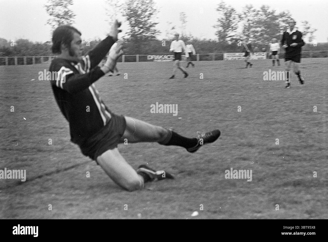 SIZO - SSS Football, Whizgle News, Dutch Desk, Niederlande, 1950 - 2000 auf 31-08-1974. Das Bild enthält diese Themen. Die Szene fängt einen Moment auf einem grasbewachsenen Fußballfeld ein, wo die Atmosphäre voller Action ist. Im Vordergrund sieht man einen Spieler, der in einem dunklen Trikot mit hellen Streifen gekleidet ist, zu Boden fallen, wobei eine Hand ausstreckt, als ob er versuchen würde, sich zu stabilisieren. Die Bewegung deutet auf ein intensives Spiel hin, möglicherweise mit einem Tackle oder einem plötzlichen Verlust des Gleichgewichts. Die Shorts des Spielers haben einen tiefen Schatten und werden durch lange, hoch gezogene Socken kontrastiert, was ein auffälliges Erscheinungsbild schafft. Gras Stockfoto