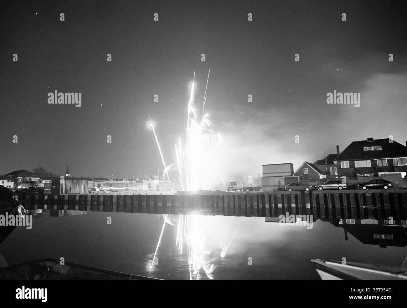 Feuerwerk in Hillegom Hillegom, Whizgle News, Dutch Desk, Niederlande, 1950 - 2000 am 20-03-1996. Diese Themen sind in der Abbildung dargestellt. In einer ruhigen nächtlichen Umgebung spiegelt das Wasser die Brillanz des Feuerwerks wider, das in Bögen platzt. Der Himmel ist dunkel, umgeben von einem tiefen Schwarz, das einen dramatischen Kontrast zu den weiß- und Grautönen des beleuchteten Feuerwerks bildet. Jede Explosion sendet schimmernde Lichtspuren, die nach unten kaskadieren und ein Gefühl von Bewegung und Energie erzeugen. Die umliegende Landschaft ist meist von der feurigen Darstellung umgeben. Bauten entlang der Küste – Häuser und p Stockfoto